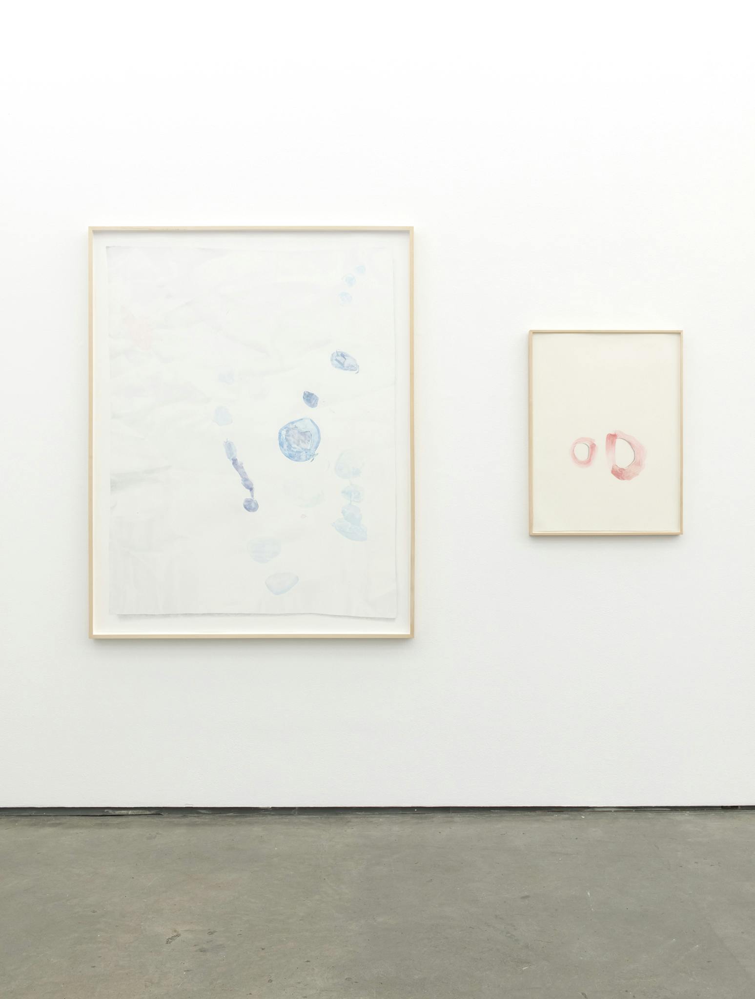 Two framed drawings on paper hanging on a gallery wall. The one to the right side is larger with blue markings, and the one on the right side is smaller with pink markings.