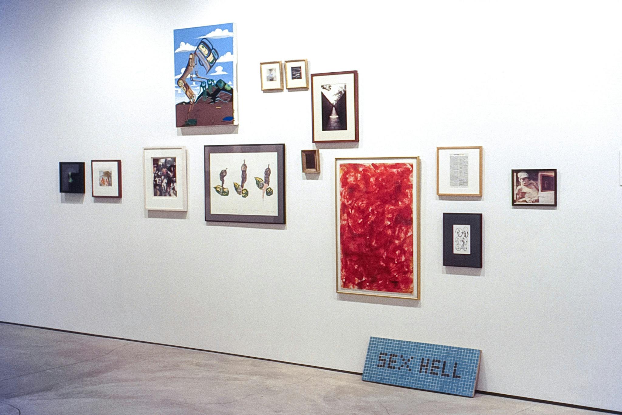 13  artworks are mounted on a white wall in a gallery, all but one are framed. At the base of the wall on the floor, there is a tile mosaic that reads "Sex Hell" in red letters on a blue background. 