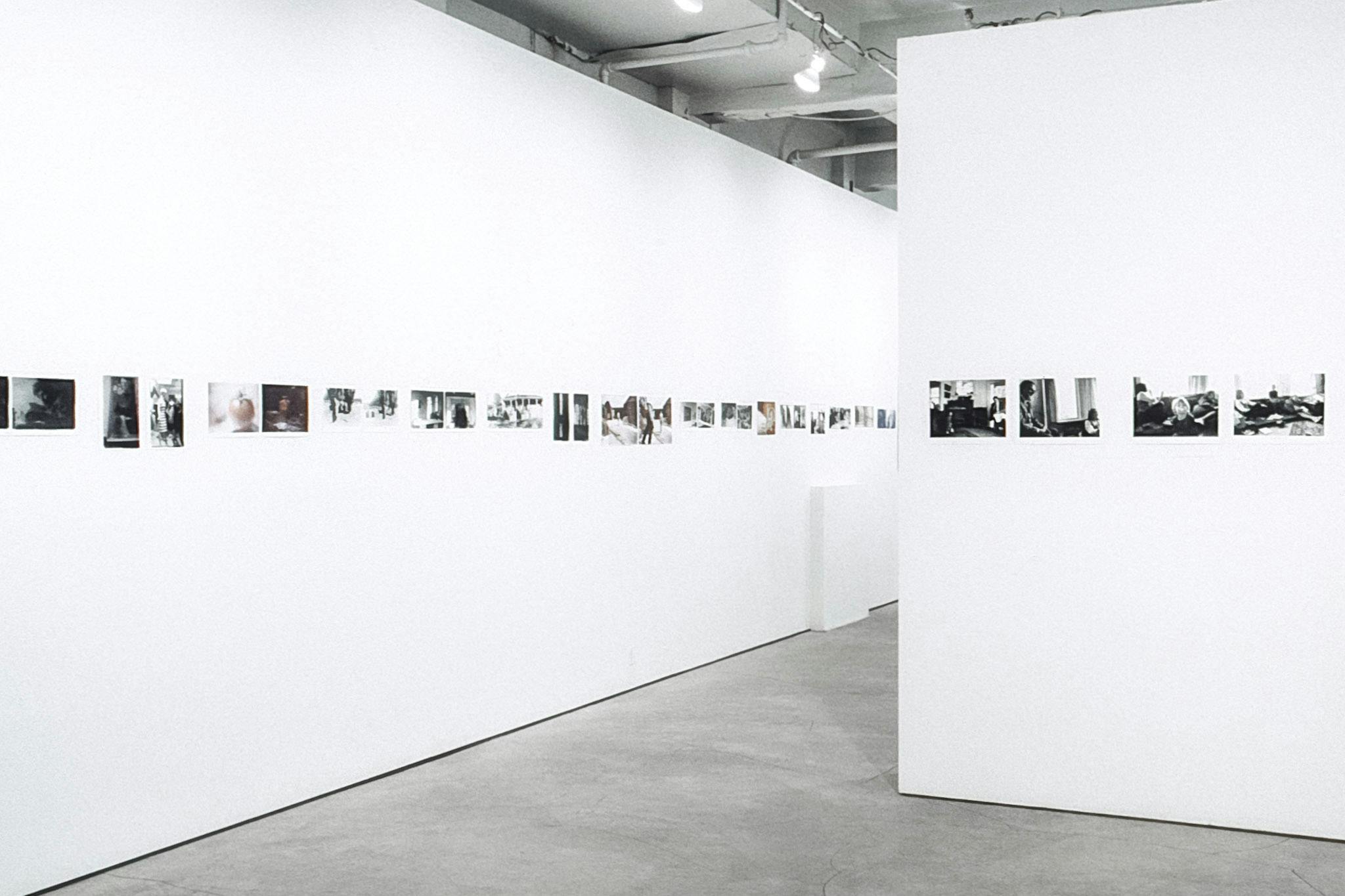 The view of a gallery space, interrupted by a wall creating a median. Across the walls, there are several colour and black and white photos arranged in a line. The photos vary in size and orientation.