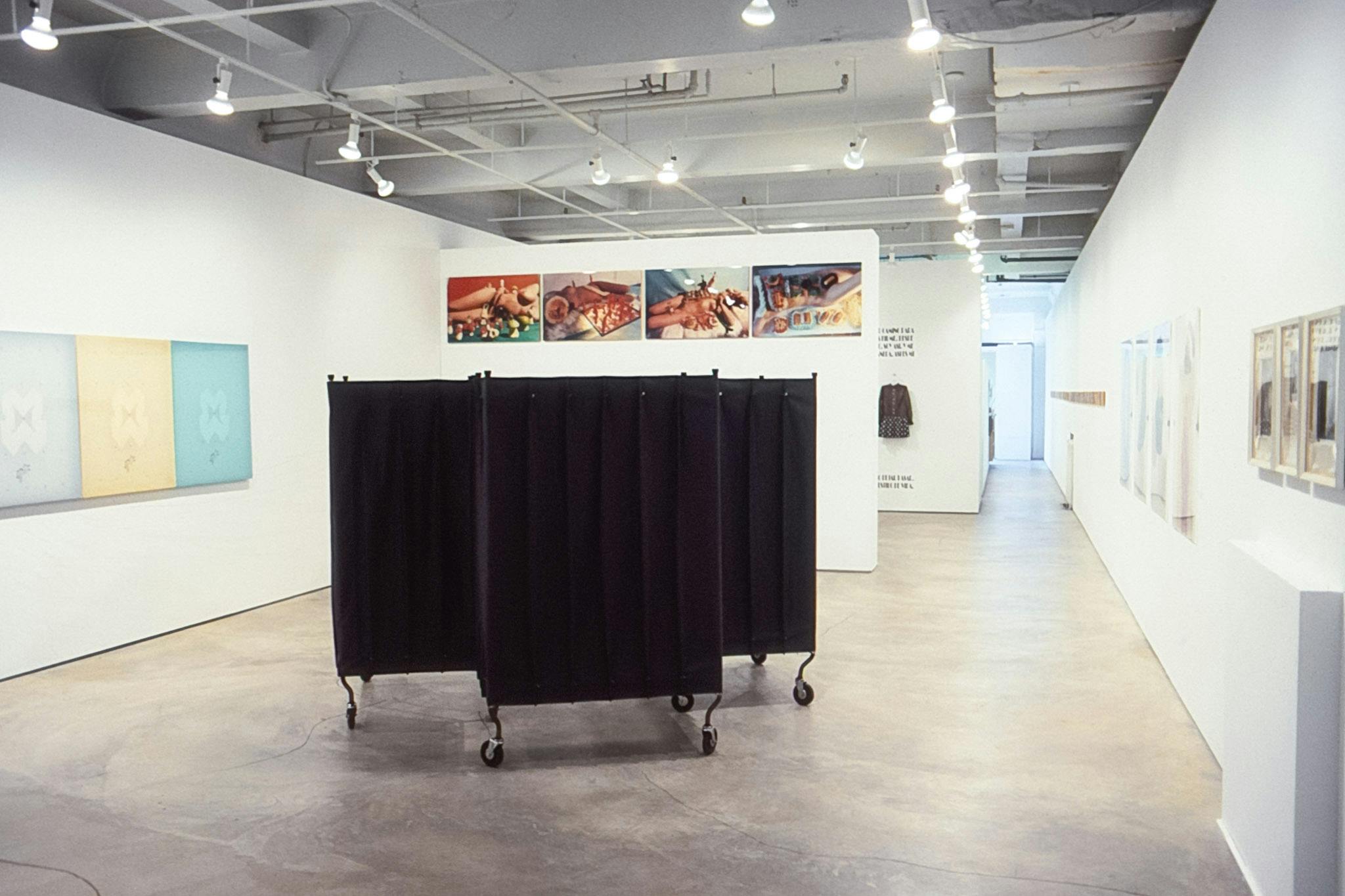 Various artworks are installed in the gallery. A pop-up changing room is placed in the middle of the floor, and a series of four photographs of a headless mannequin is mounted on a wall.