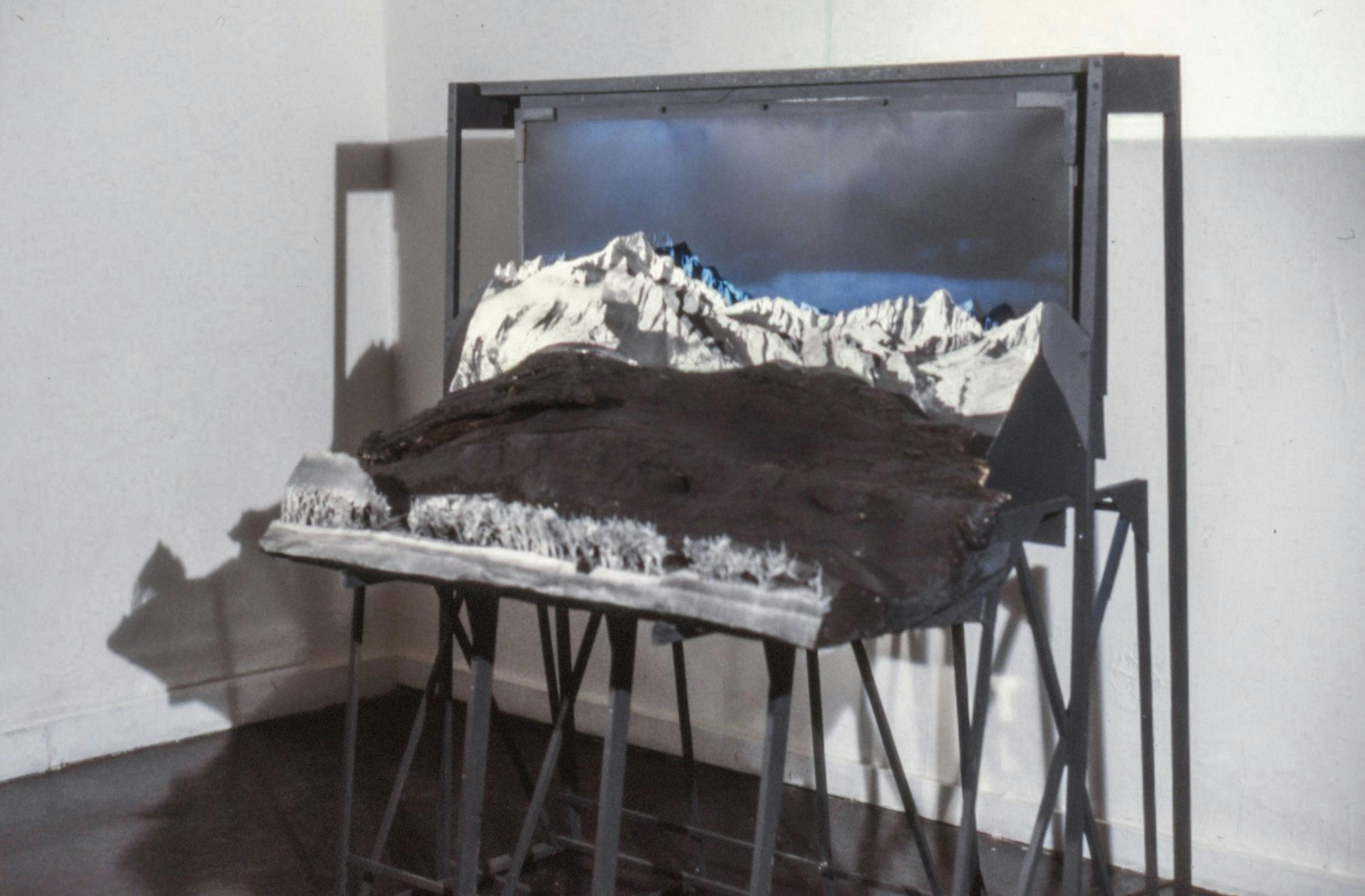 In the corner of a gallery, a large photo set rests on a black wooden stand. The set has a dark blue sky background, layered mountain forms, a black textured surface, and white trees.