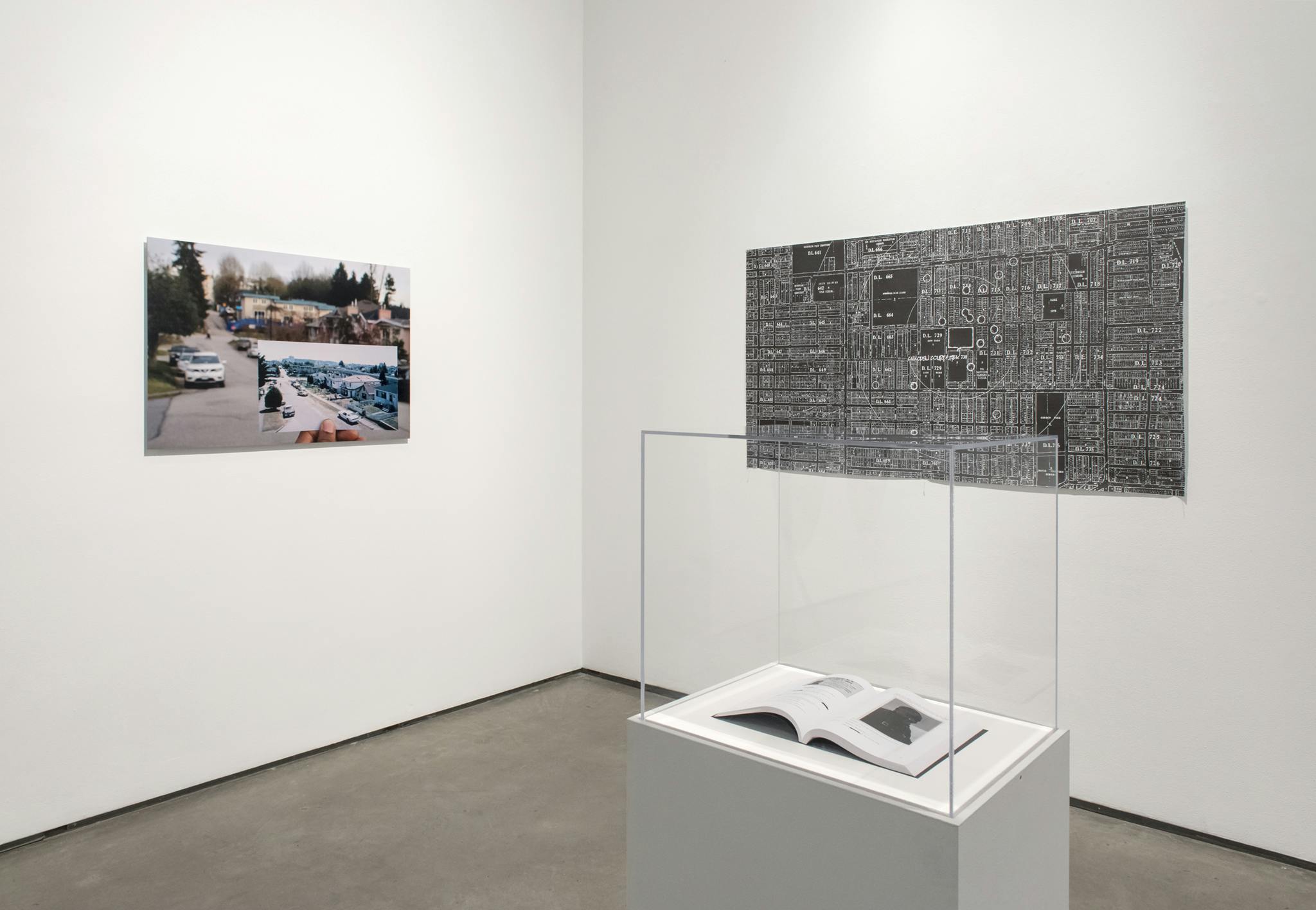A corner wall of a gallery space with a photograph and a print of city subdivisions hanging on each side of the wall. A vitrine with an open book sits to the right. 