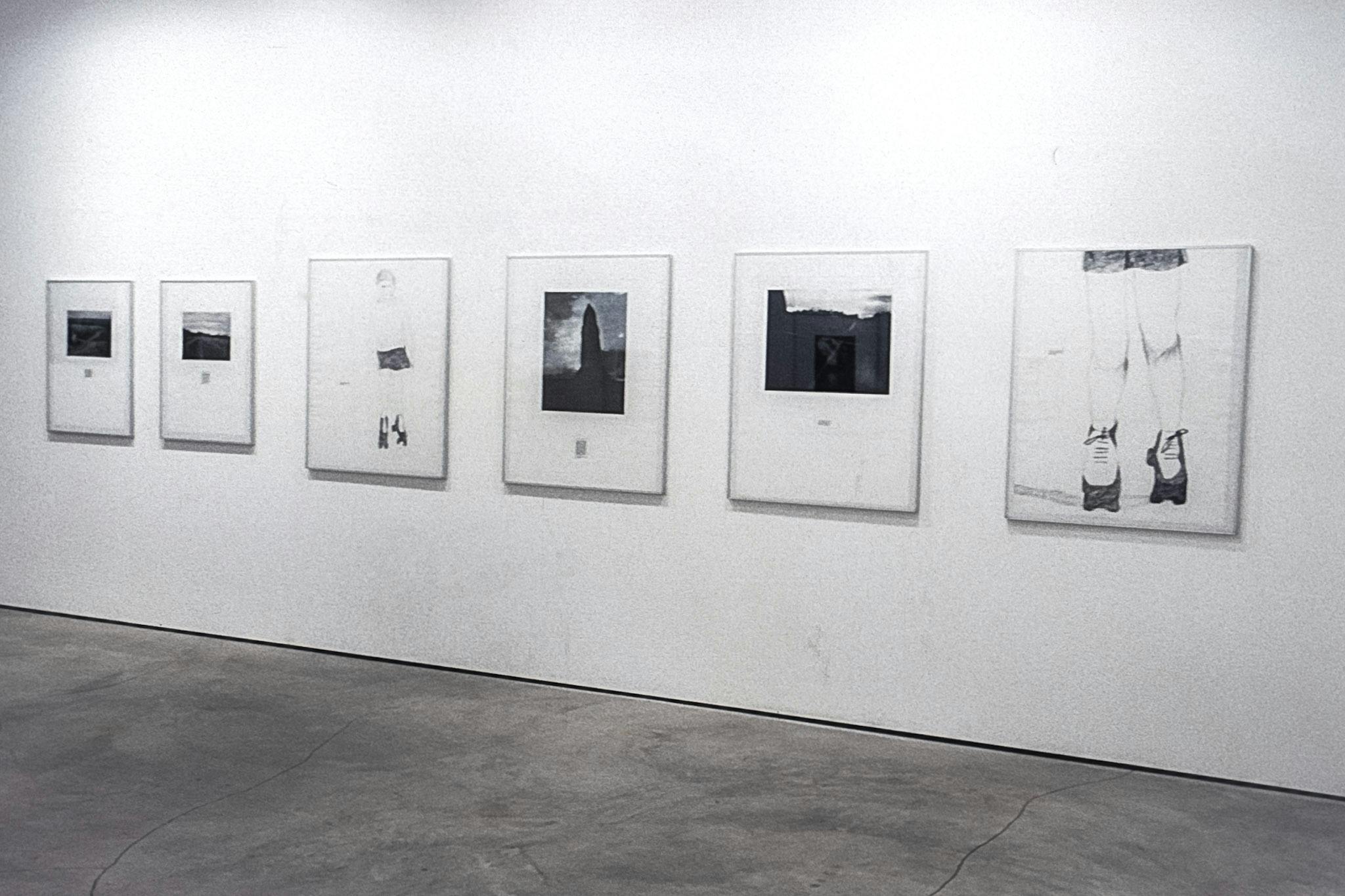A gallery space with work on the white walls. Most of the works visible are mostly-white illustrations in metal frames. In the background, a partial black and white photo of a stadium can be seen.