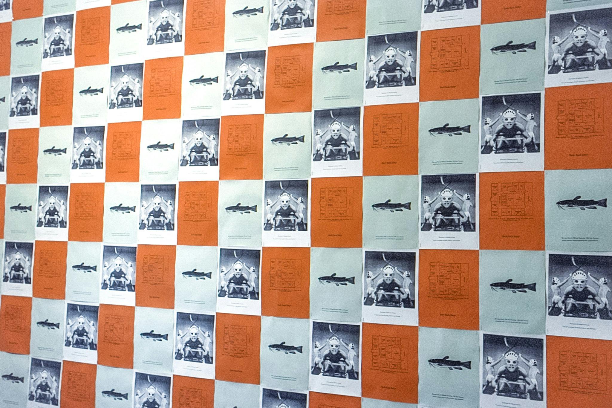 A gallery wall covered in 3 repeating images. One is a blueprint on orange paper, one is an illustration of a catfish on green paper and the other is a still from the film "The Making of the Monster."