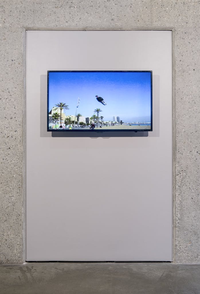 A monitor on a gallery wall plays a single-channel video piece. A person wearing a black jacket flies above a beach in the blue sky as if they were a kite.
