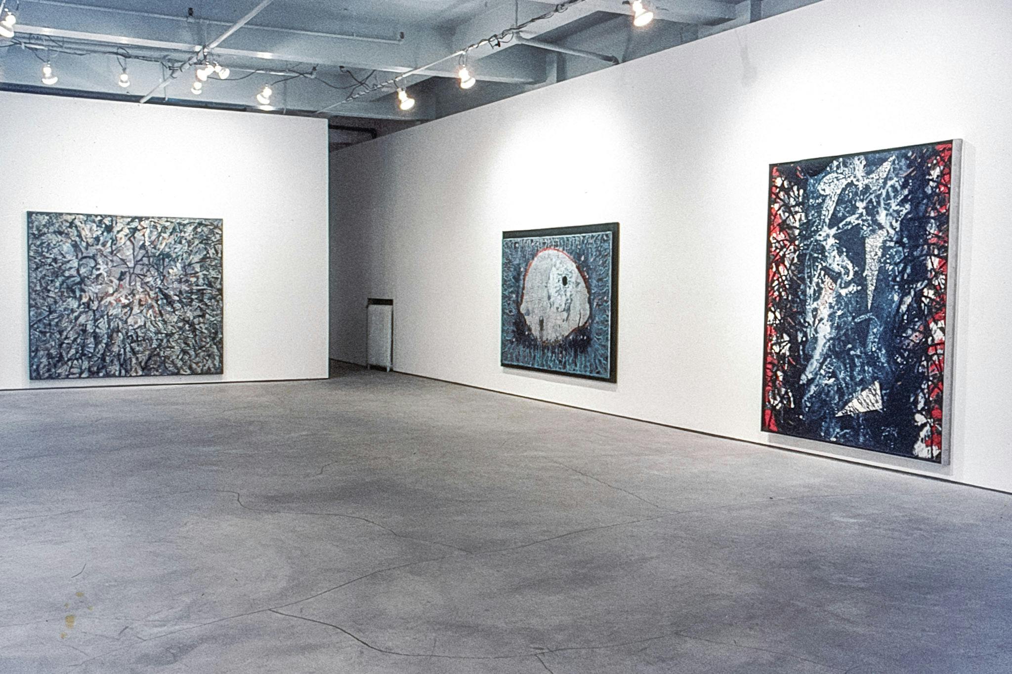 3 paintings in a gallery. The paintings are all big but vary slightly in shape. They show different forms created through layers of paint and materials, primarily using red, black, white, and blue.