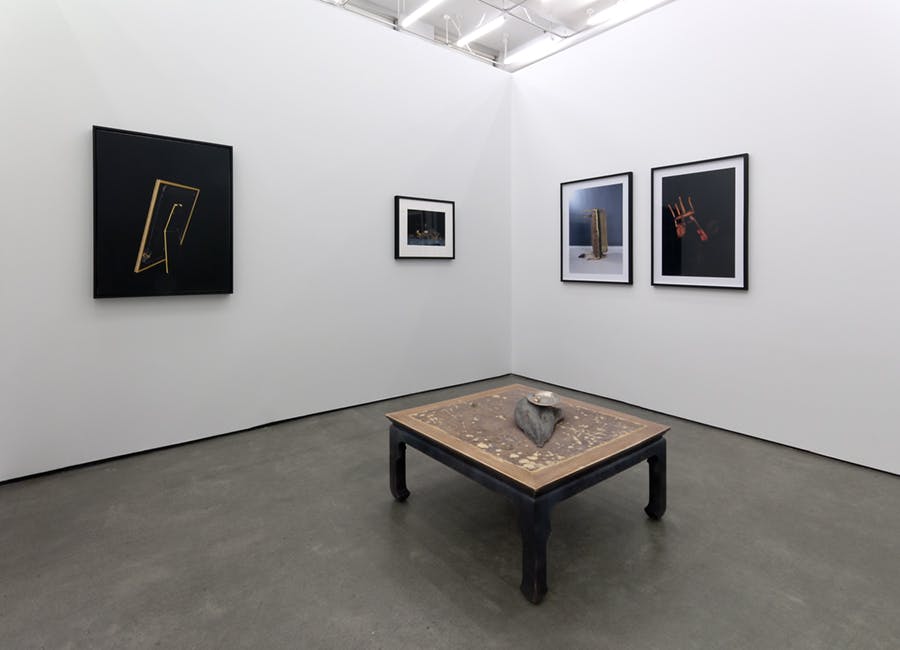 Four framed photographs hang on either side of a corner wall in a gallery. A small table with a brown top and black legs sits in the middle. A smaller object sits on top of the table.