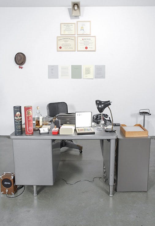 An installation image of a work by Daniel Olson. A steel-made grey working desk is placed in front of the wall. The desk is covered with various objects, including a typewriter, a desk lamp, and a bottle of rum.
