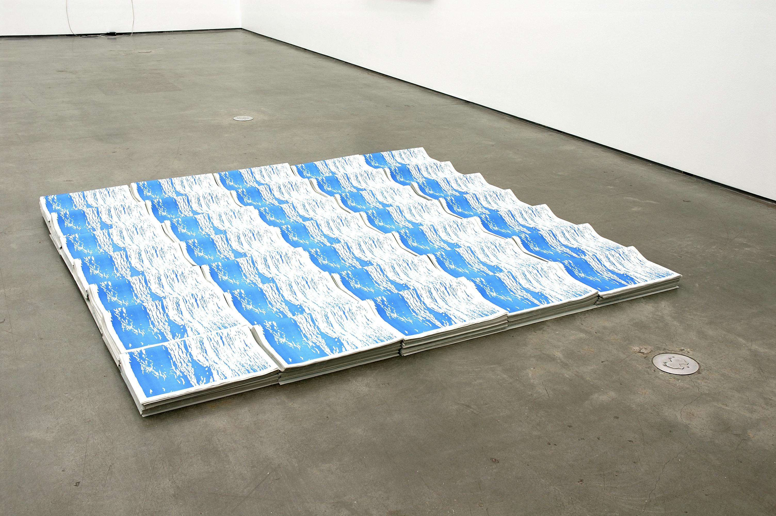 7 times 5 stacks of brochures are placed on the grey gallery floor. The brochures are made of newsprint paper. Repetition of both their shape and blue and white cover image resembles water waves. 
