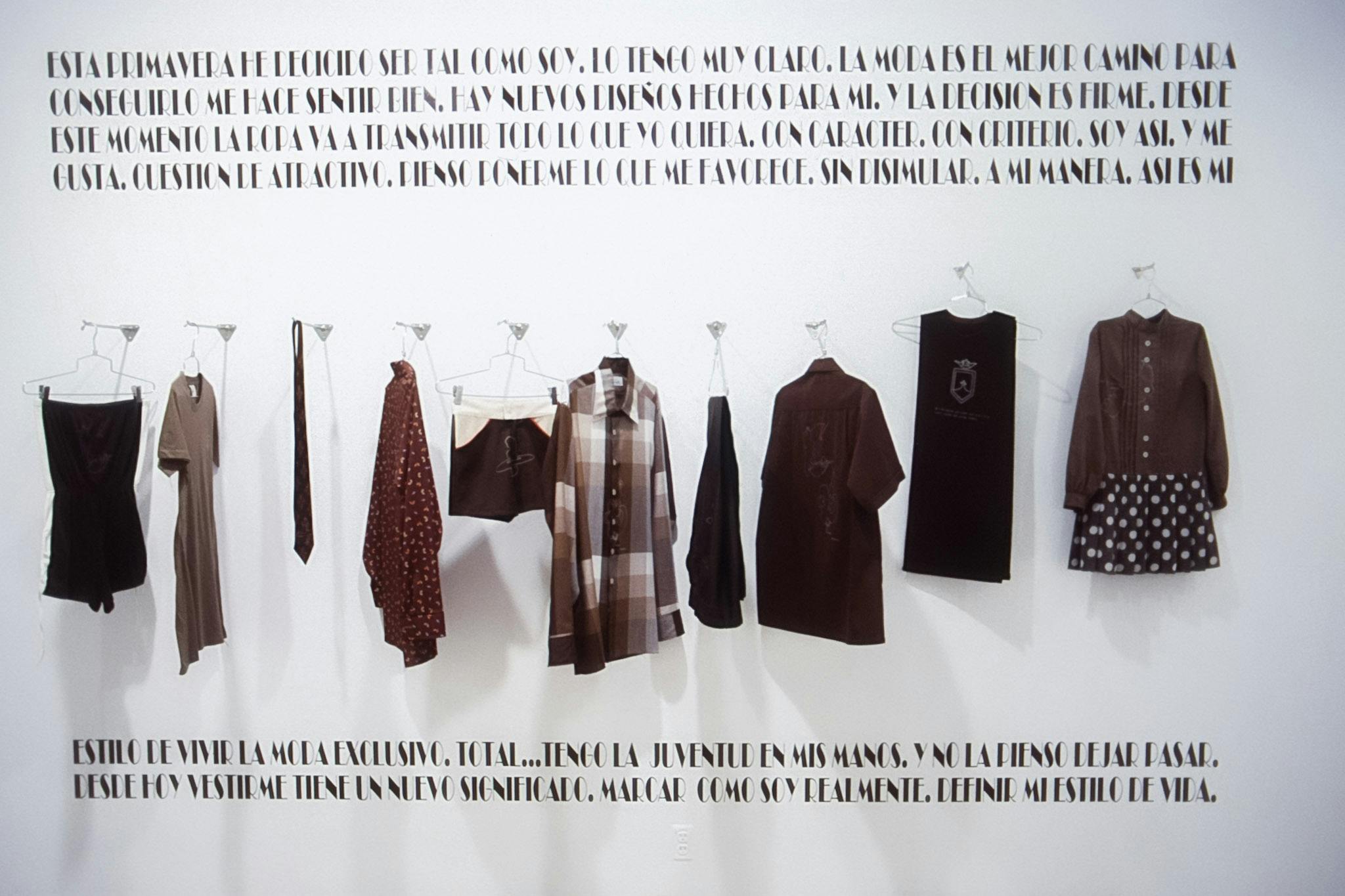 A front view of an installation artwork. Ten brown colored clothes are hung on the white gallery wall. There are Spanish texts written both above and below the clothes.  