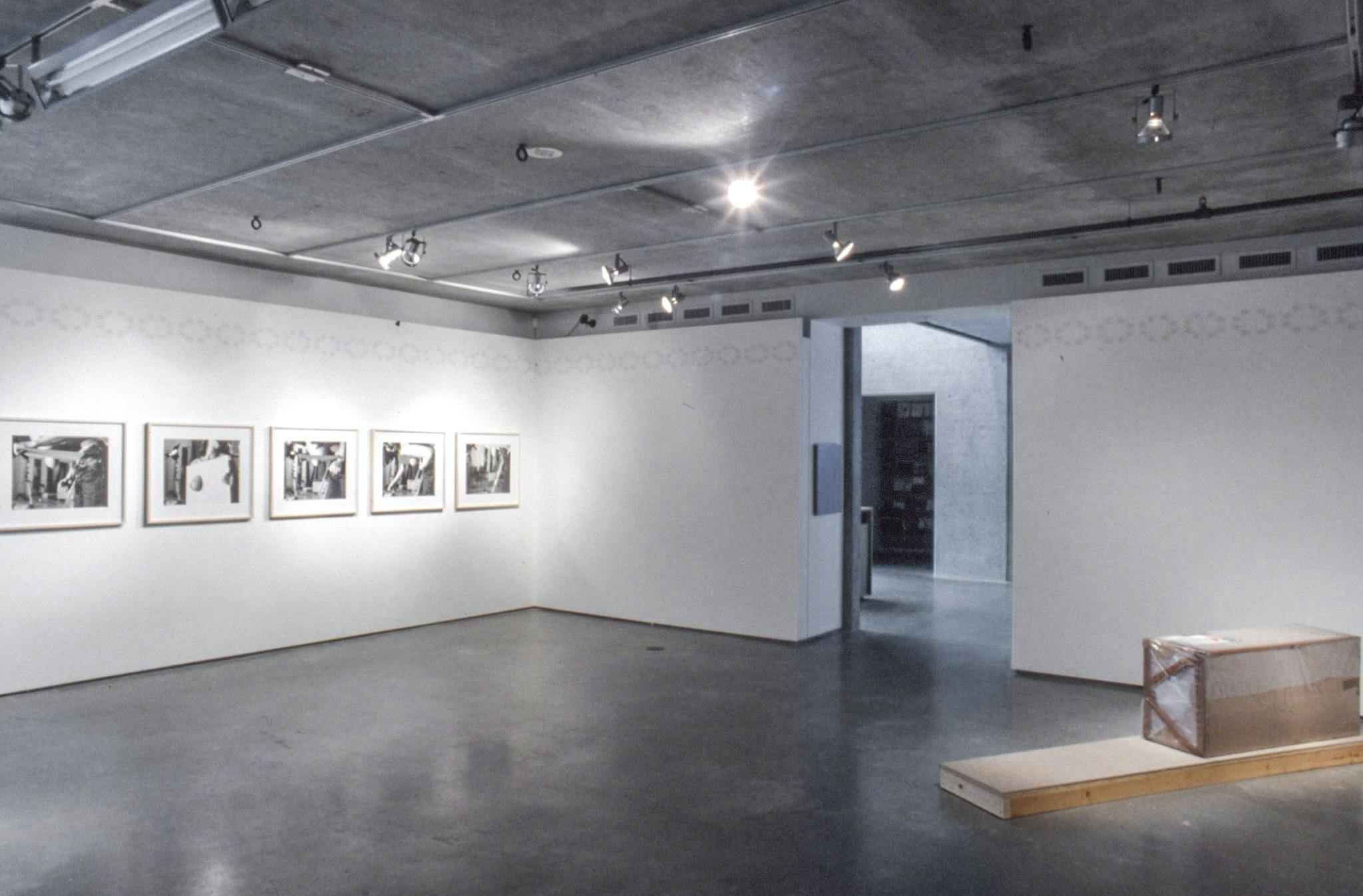 Several artworks are installed in a gallery space. Five framed black and white photographs are mounted on the left side wall. A wood sculpture is installed in a corner of the room. 