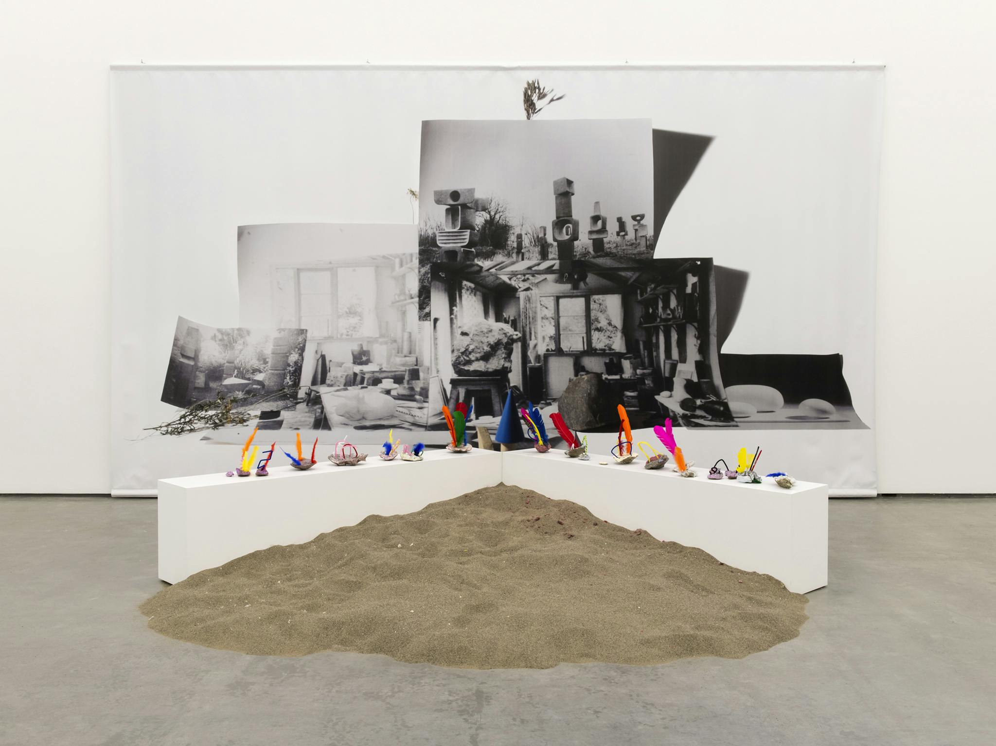 Sand is piled against a V-shaped plinth on a gallery floor. Lining the top of the plinth are small colourful sculptures. Behind a large black & white vinyl print of a photo collage hangs on the wall. 