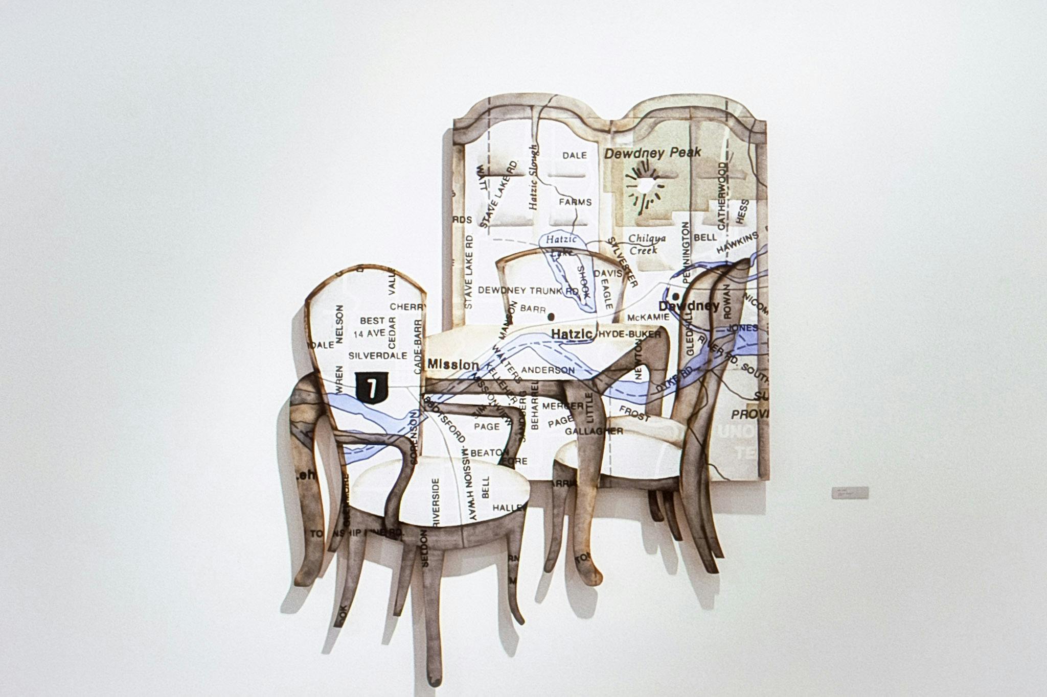 A painting collage on a white wall. The painting shows 3 cushioned chairs and a table, textured with, and layered in front of, a map showing roads and rivers. 