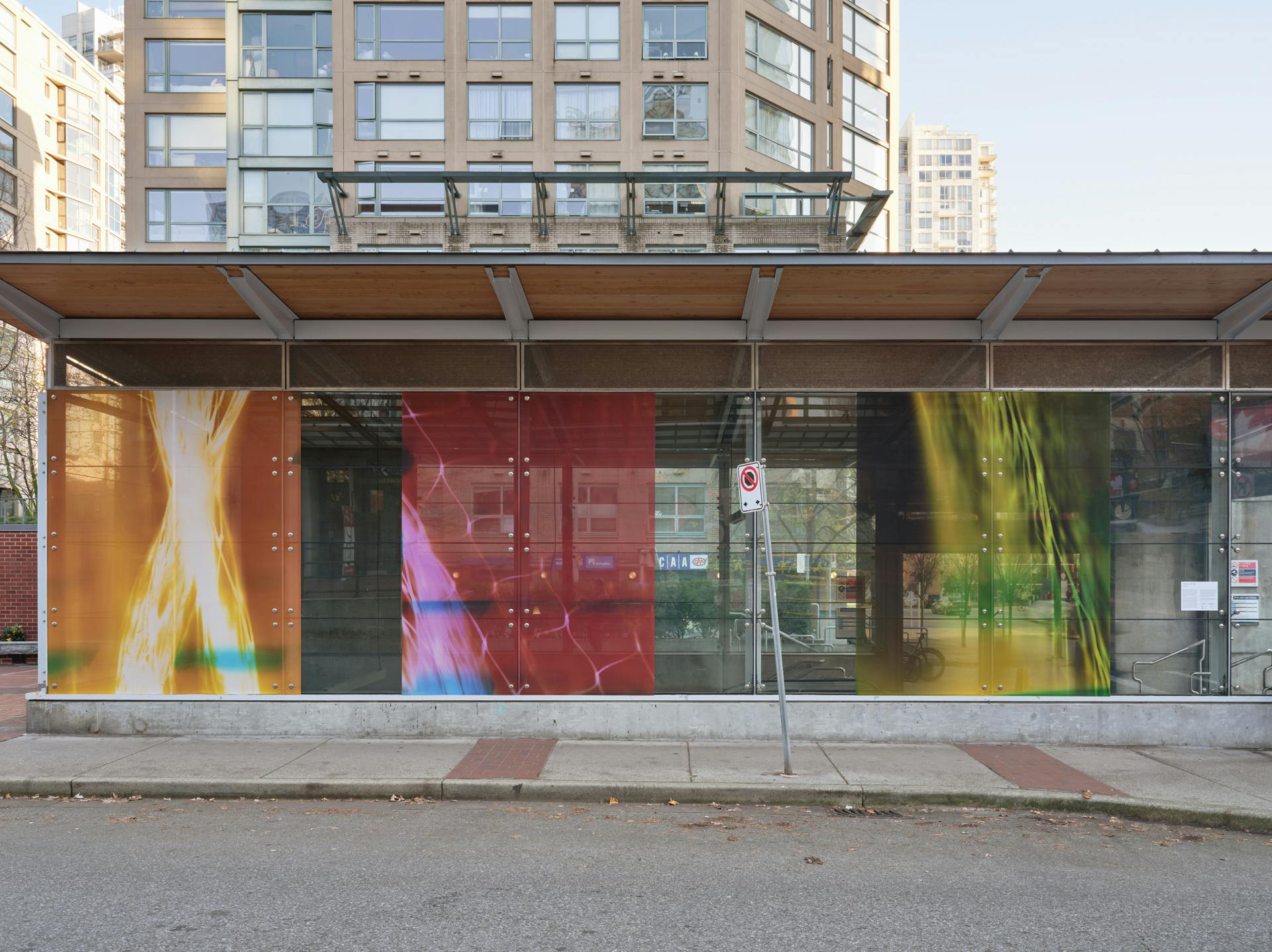 "A frontal view of a SkyTrain station with three photograms in its windows depicting mesh nylon bags against neon photographic paper. The left photogram resembles an orange X. The centre is red and pink. The right photogram is green and yellow "