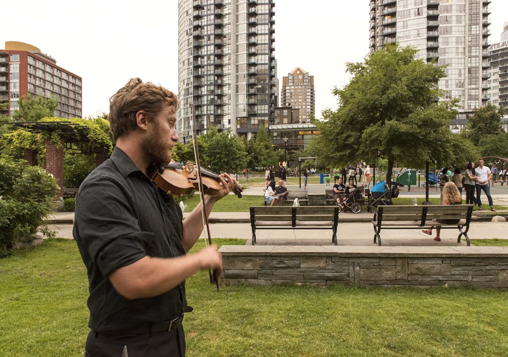 A person dressed in black plays the violin outdoors in a park. There are buildings, trees and benches in the background. 