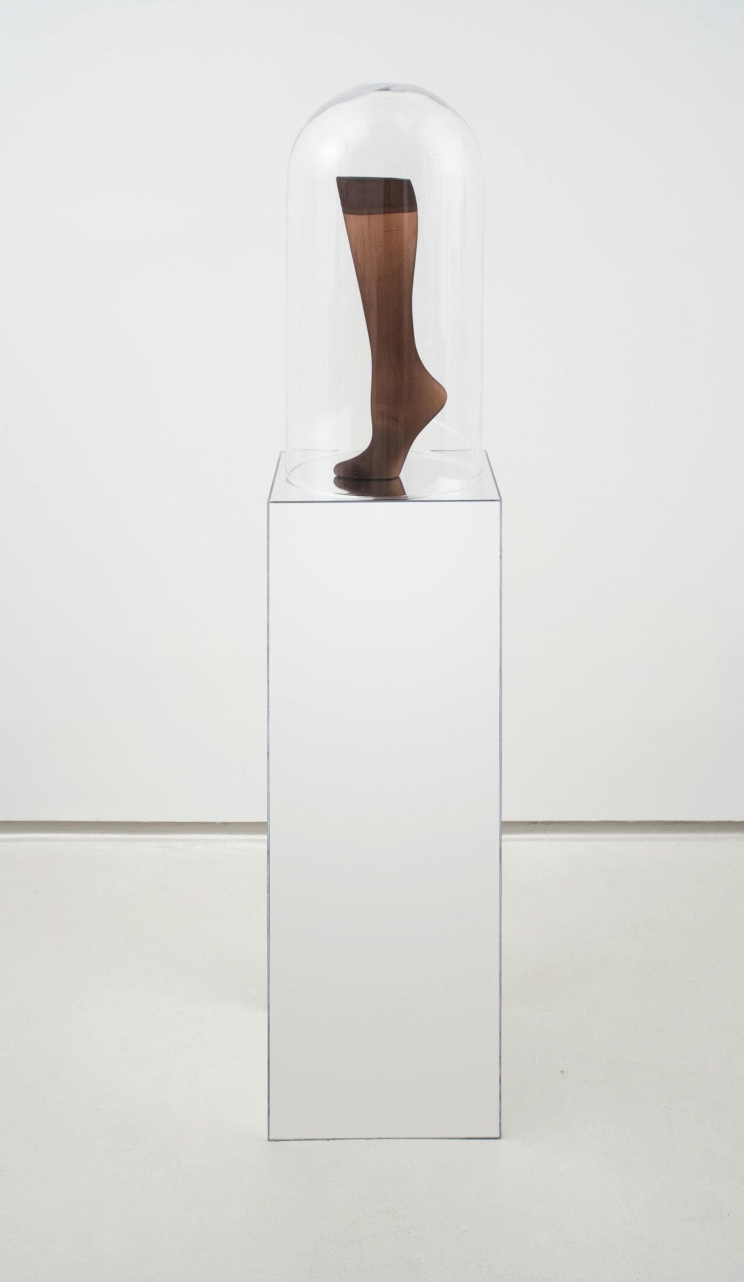 A sculpture made of a mannequin foot in brown stockings covered by a glass bell jar sitting on a mirrored plinth.