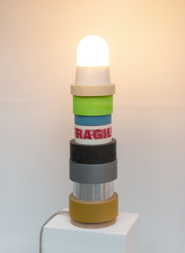 A sculpture made of various rolls of duct tape are stacked one on top of the other. A dome-shaped incandescent lightbulb shines from the top. 