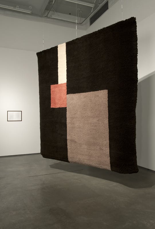 A large, hand-knotted carpet is installed in a gallery space. Hanging from the ceiling, it is square-shaped and mostly black in colour except for 3 rectangles in brown, pink, and white in the middle.
