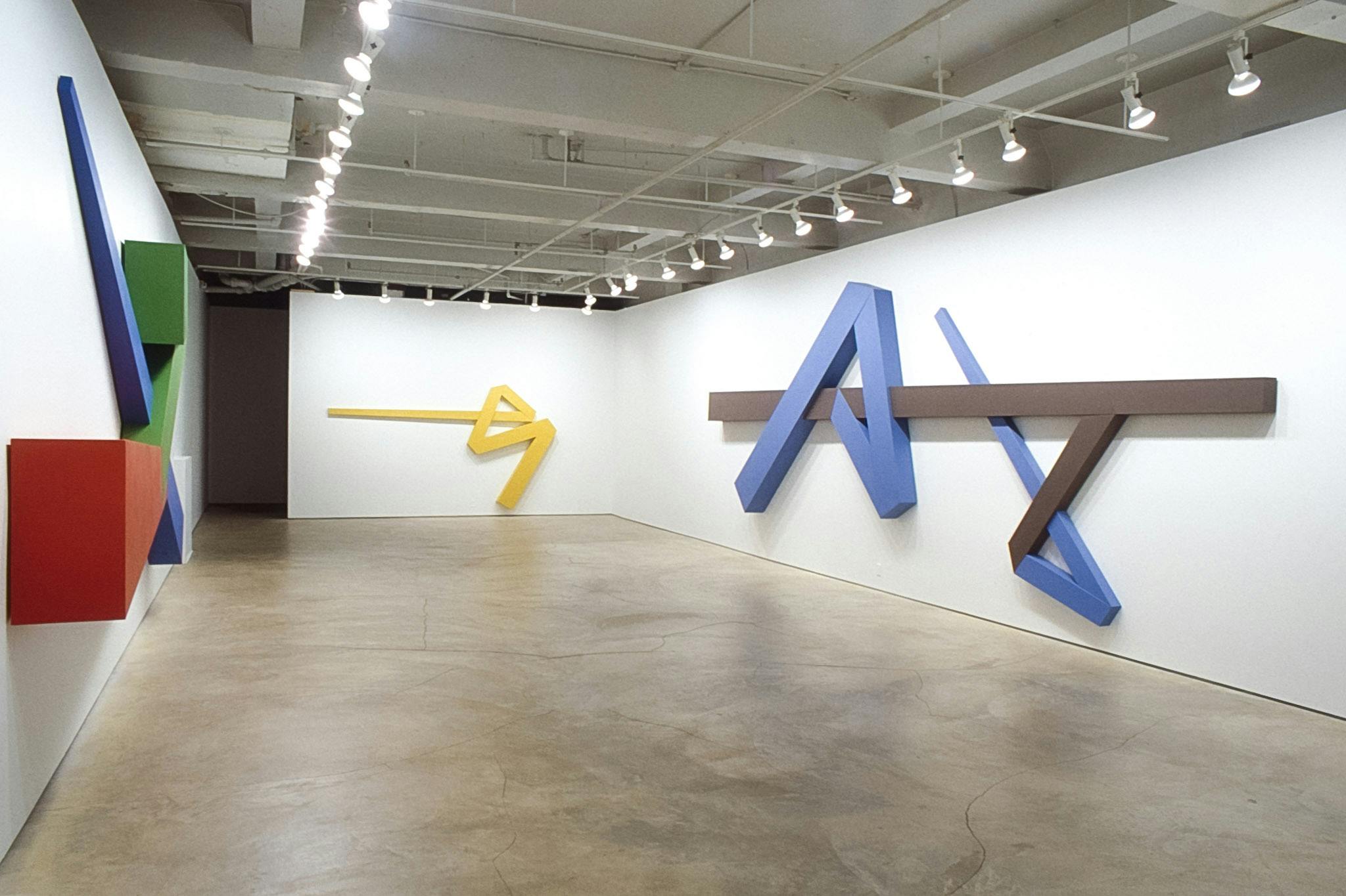 Three large-sized geometric sculptures are mounted on the gallery walls. The one on the right is composed of blue and dark-brown colored shapes. The middle piece is yellow. 