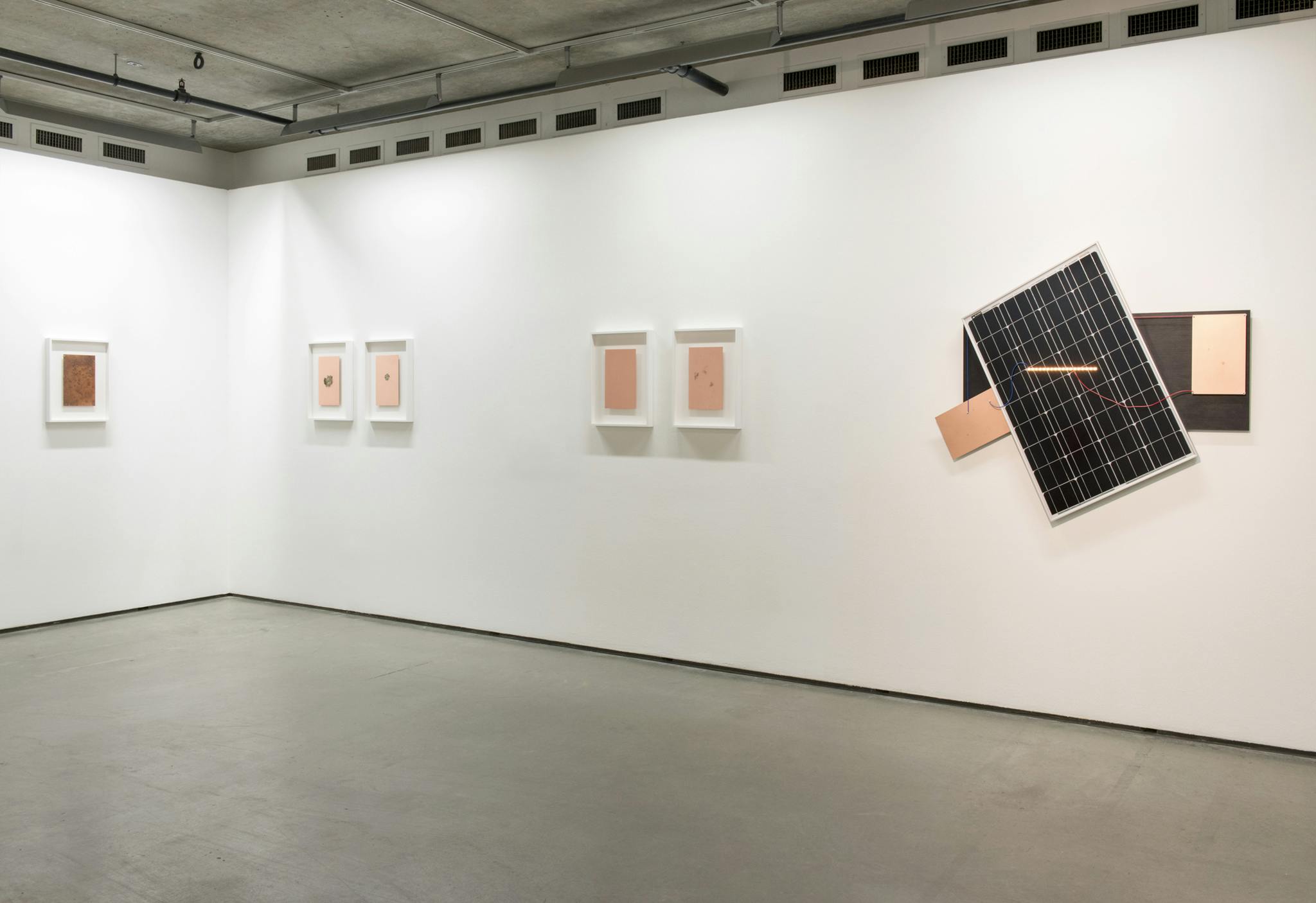Framed copper plates are mounted on two gallery walls. A large flat sculpture made of a solar panel, a bar of small orange LED lights, electrical wires, and copper plates are also mounted on a wall.