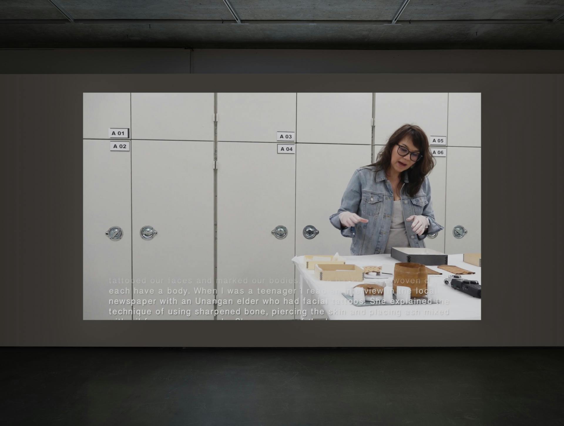Video by Tanya Lukin Linklater projected on a wall in a dark room. Video depicts Lukin Linklater wearing white gloves in front of storage lockers, speaking at a table with archival boxes and objects.