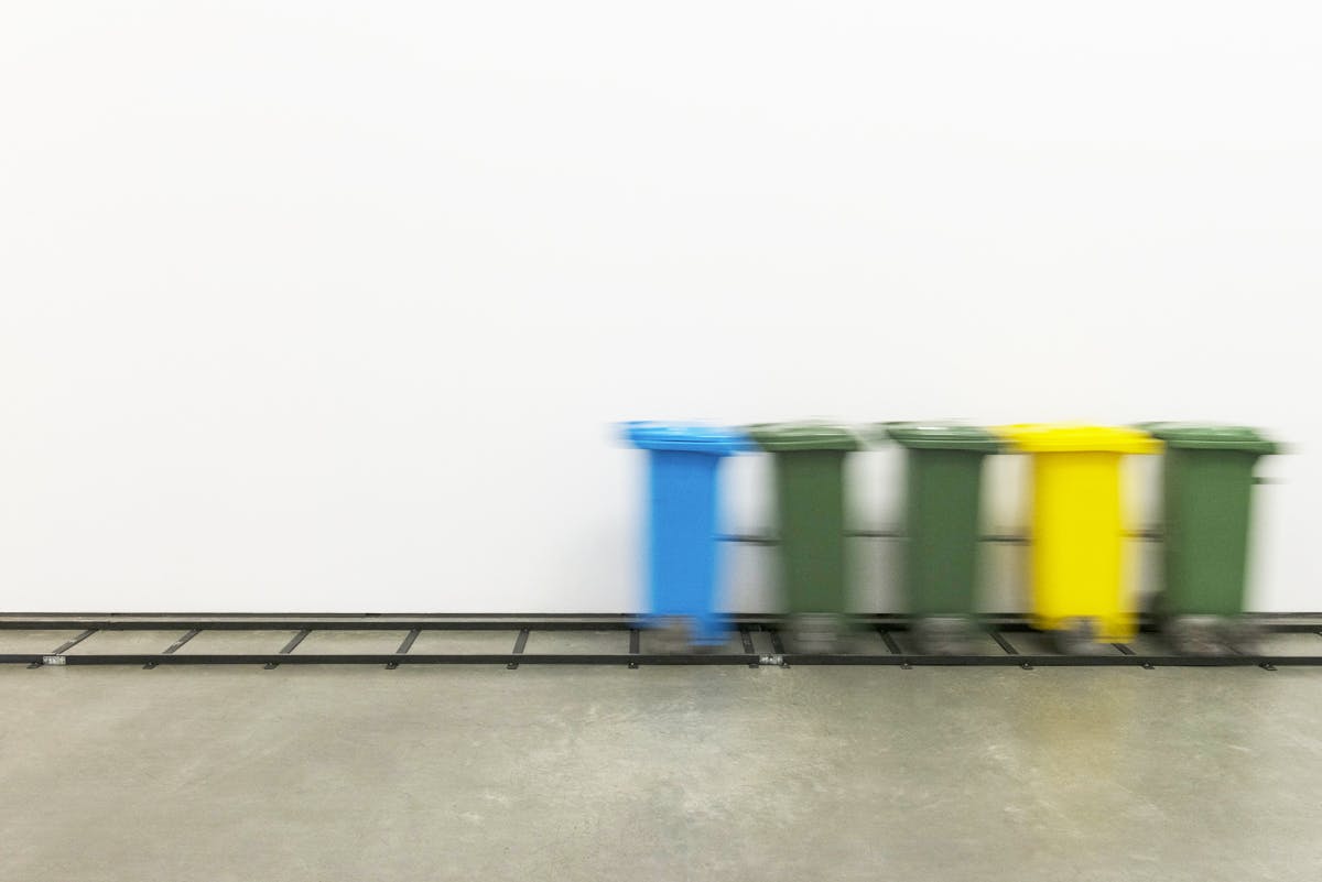 Green, yellow, and blue garbage bins appear in motion, connected to a rail built in a gallery space.