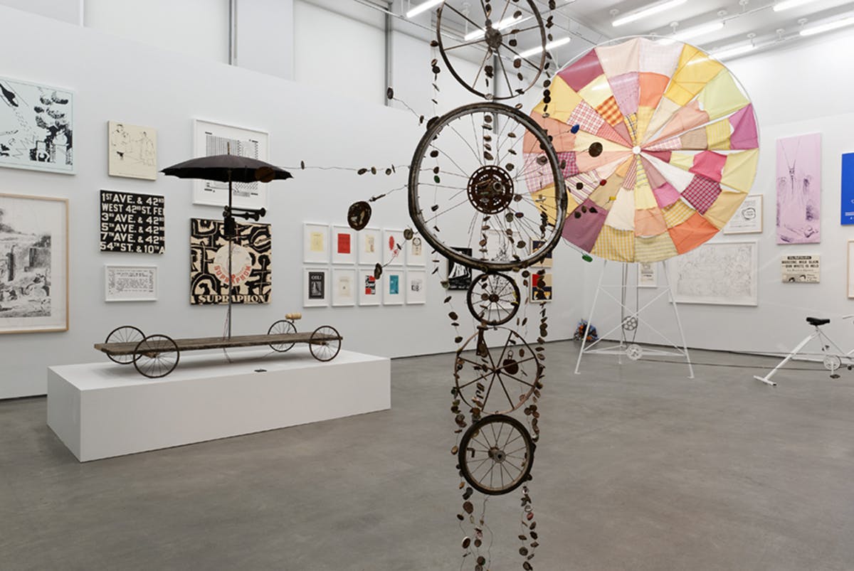 Artworks installed in a gallery. A sculpture of rusted wheels hangs from the ceiling. Behind it are large sculptures resembling a cart and a wind wheel along with many two-dimensional works on the walls.