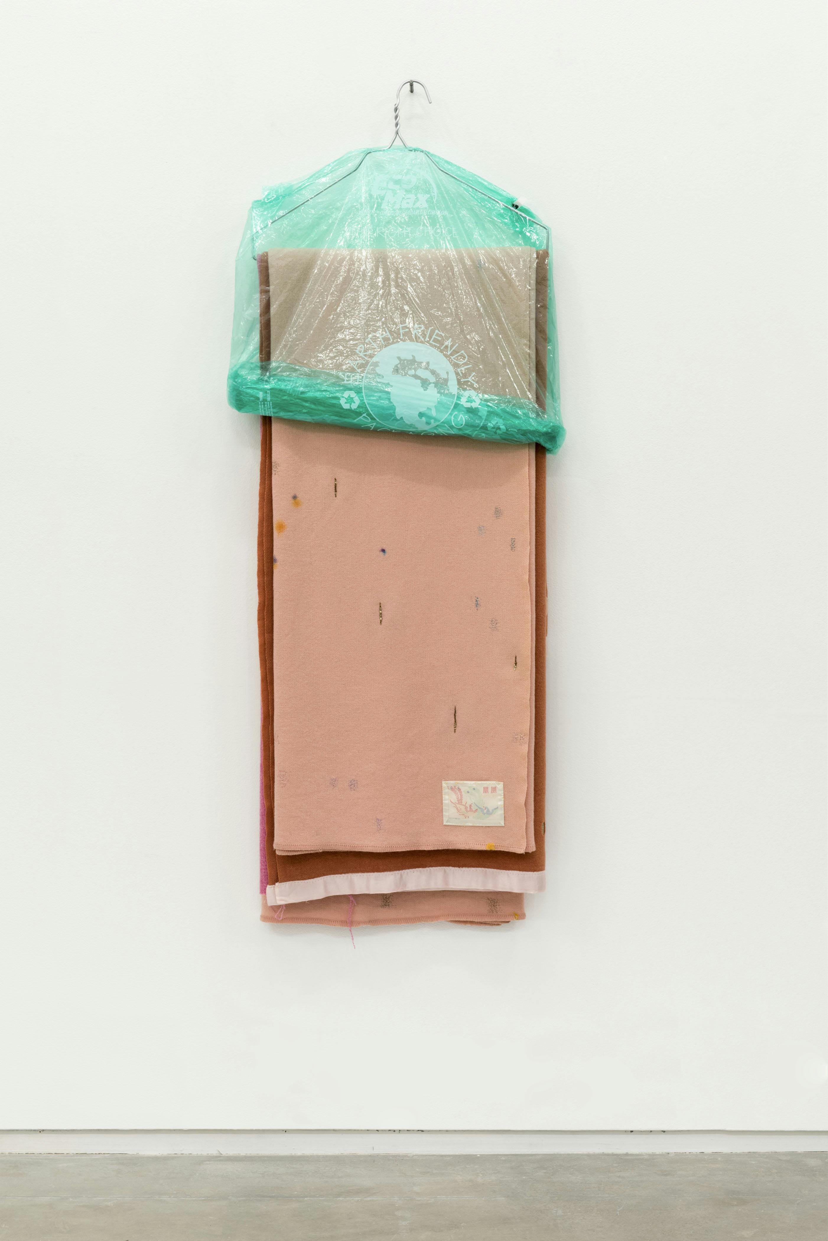 Peach and brown coloured textiles are draped on a wire-hanger which hangs on a gallery wall. A green, plastic garment bag covers half of the textile.