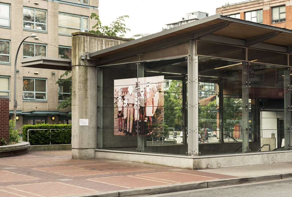 A large-scale photograph in vinyl installed on the glass exterior of Yaletown-Roundhouse Station. In the image, a family stands in front of a car. Mask-like shapes have been painted over their faces.
