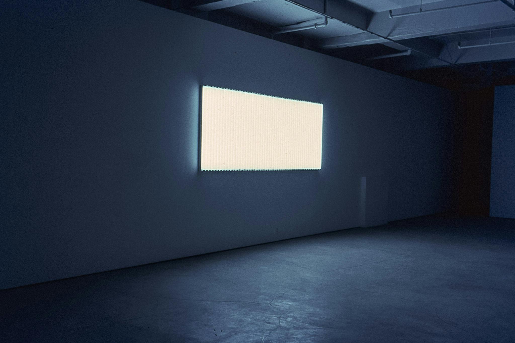 In a dark gallery space, there is a large, glowing panel mounted on the wall. The panel is a wide rectangle, with several thin strips of material running across it vertically.