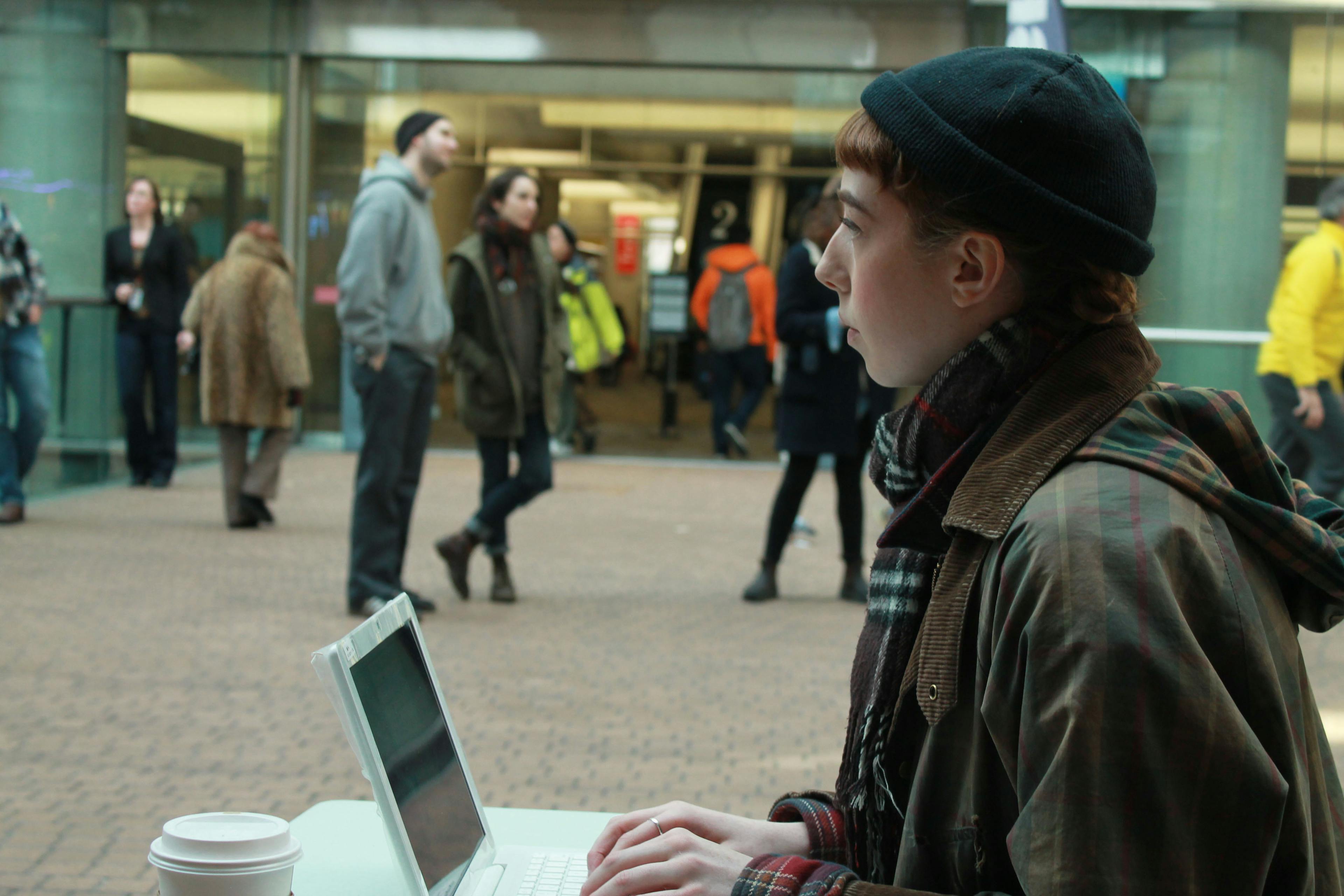 A person sits at a white desk outside an entrance to a public building. A white laptop sits on the desk. The person looks out at people entering and exiting while typing on the laptop.