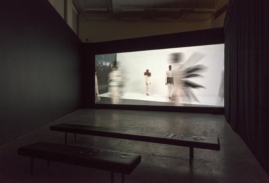 A screen covers a gallery wall beside a closed, black curtain. A video work of a performance is projected onto the screen. Several benches are placed on the floor in front. 