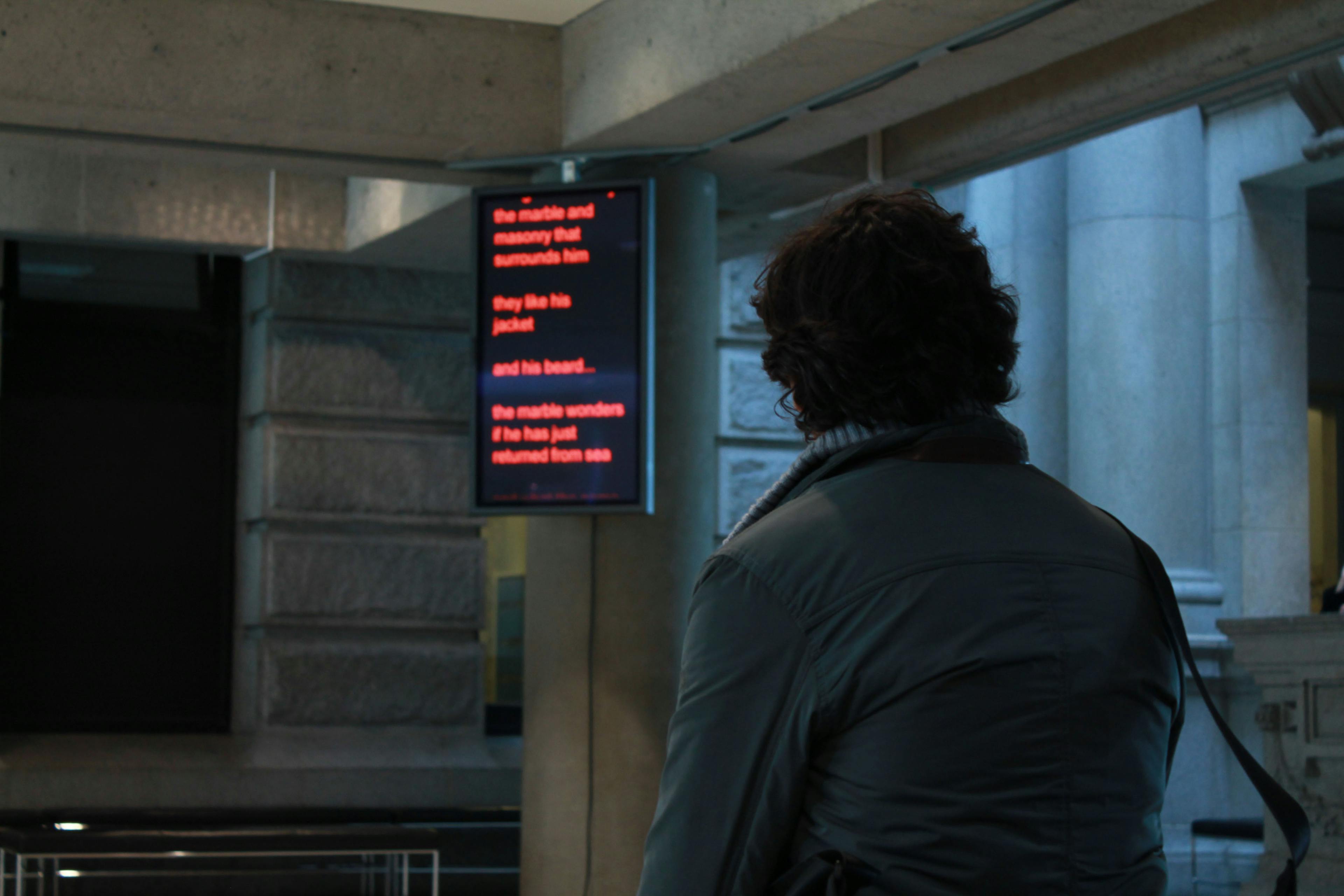 A person in a grey jacket looks at a monitor installed in an upper corner of an indoor space. The screen is black with illuminated text in red.