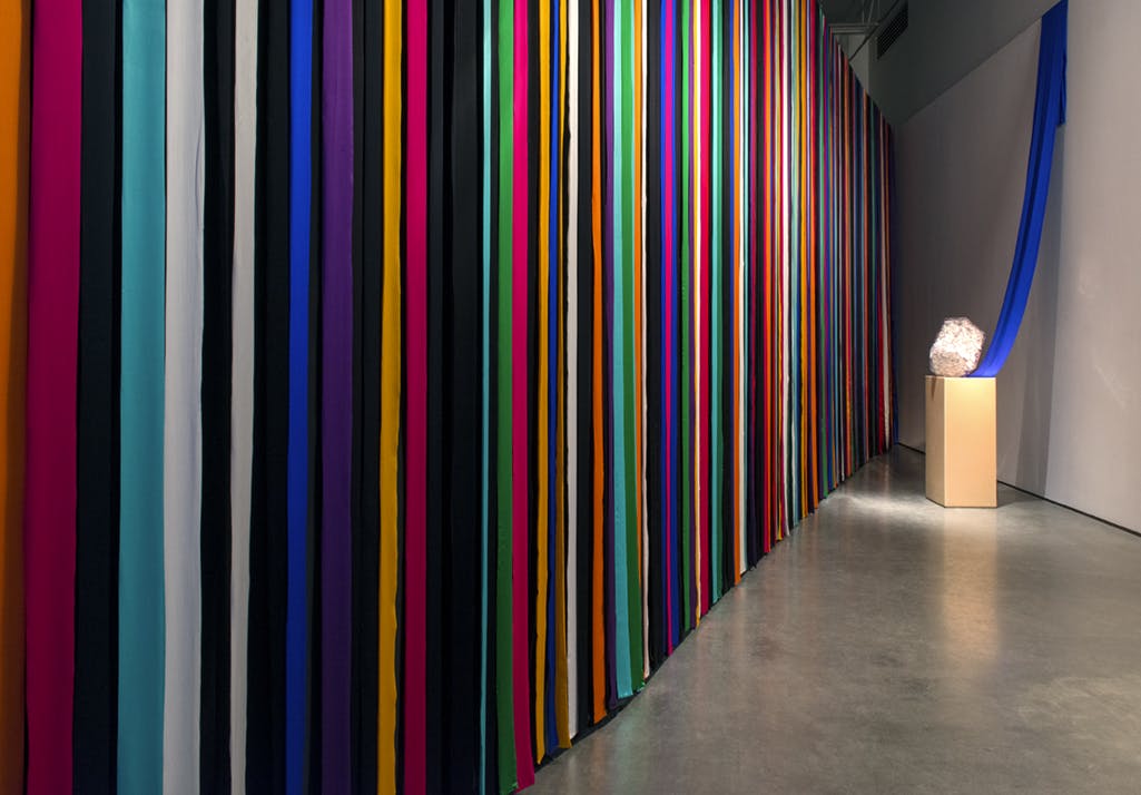 A striped multicoloured curtain hangs in a gallery. Installed in front of the curtain is a clear, block-like sculpture of encased ceramic pieces on a plinth.