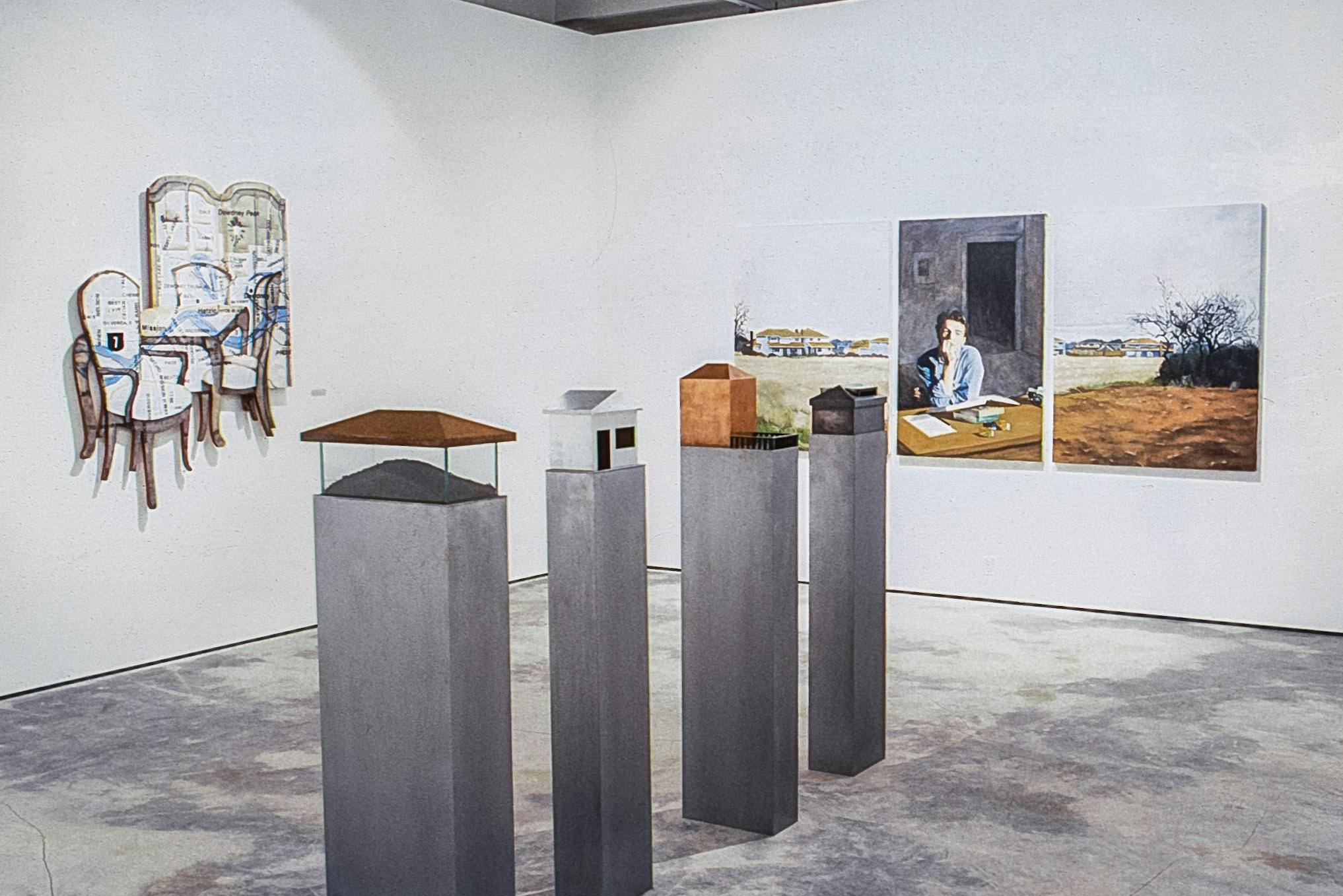 A gallery with several artworks. On one wall there are 3 paintings, on the other there is a painting collage. IN the centre, 4 grey plinths show small metal house-like forms on them.