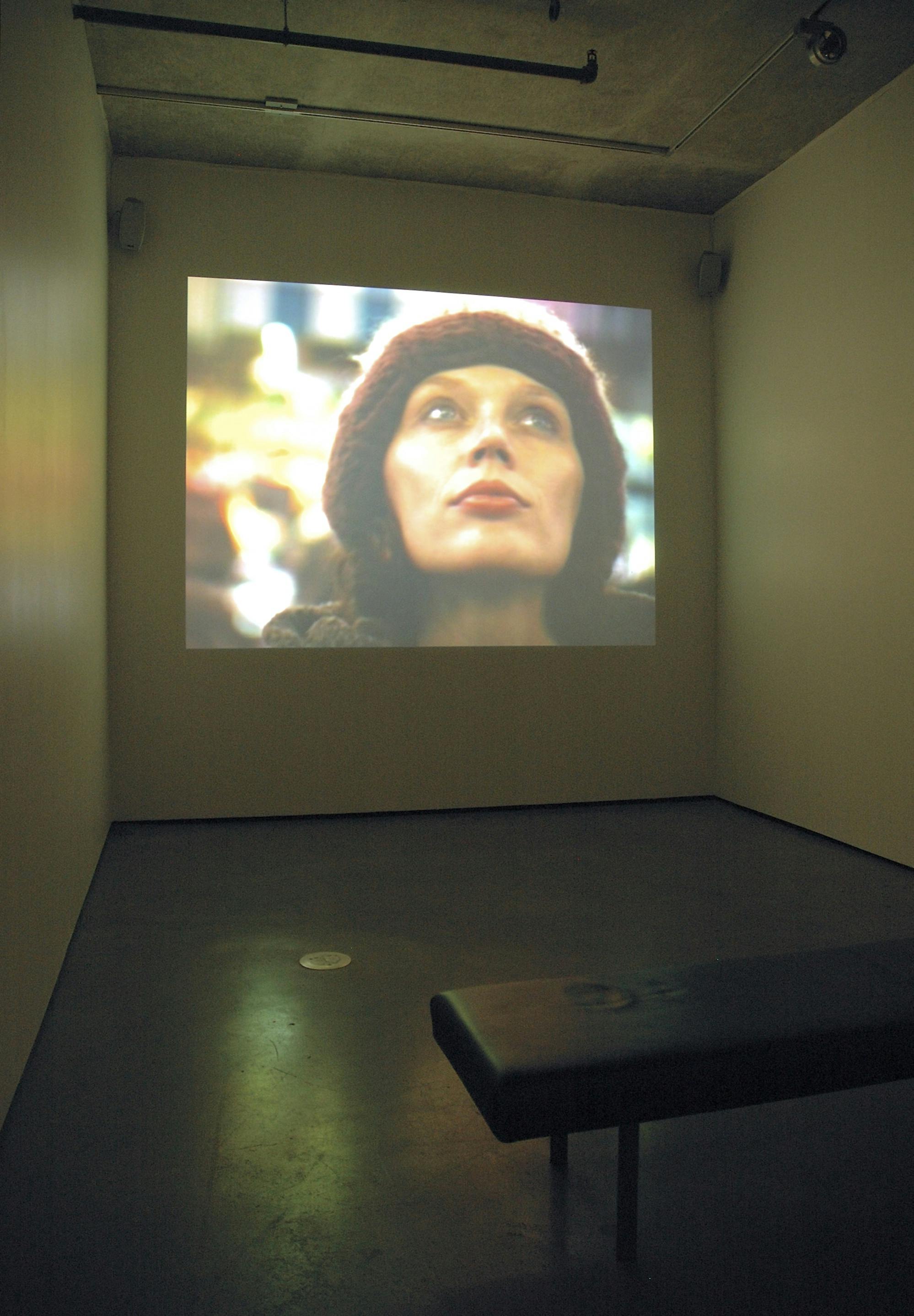 A single-channel video work is projected on one of the gallery walls. The video shows a close-up of a person’s face. The person has blue eyes and wears a red toque.