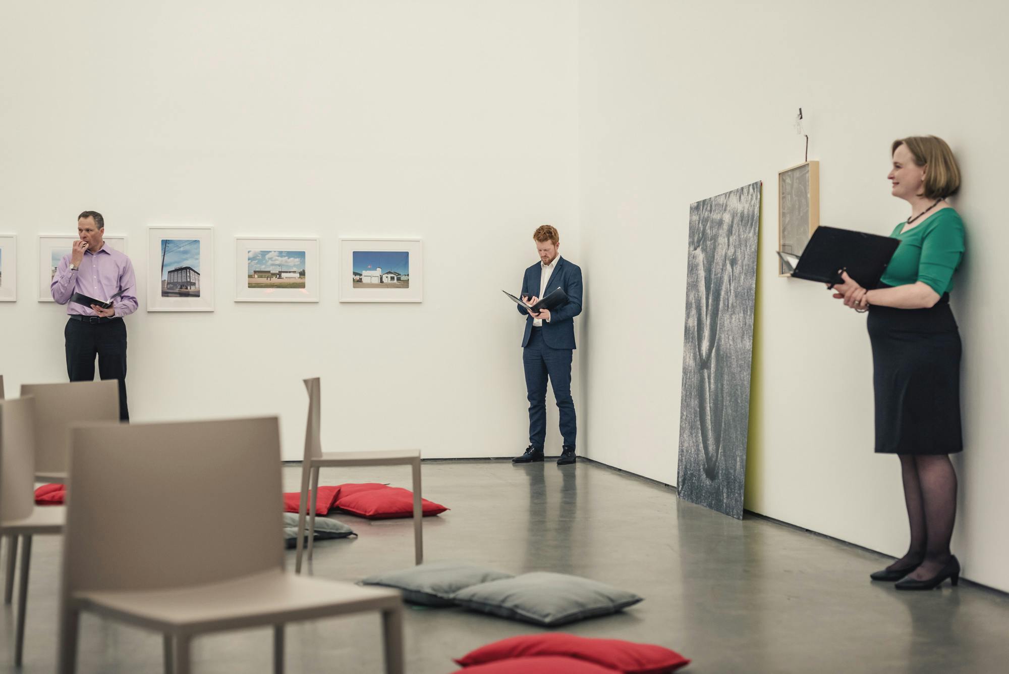 An image of people standing apart in a gallery, interspersed by artworks on the wall. They are holding books. Some chairs are placed in the middle of the gallery facing performers. 