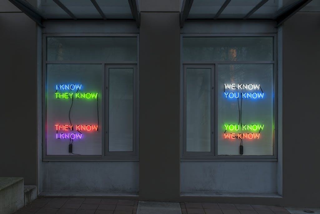 Detail image of Tim Etchells’ neon text works installed in CAG’s facade windows. Phrases in neon lights of various colours read: “I KNOW THEY KNOW,” and “WE KNOW YOU KNOW.”