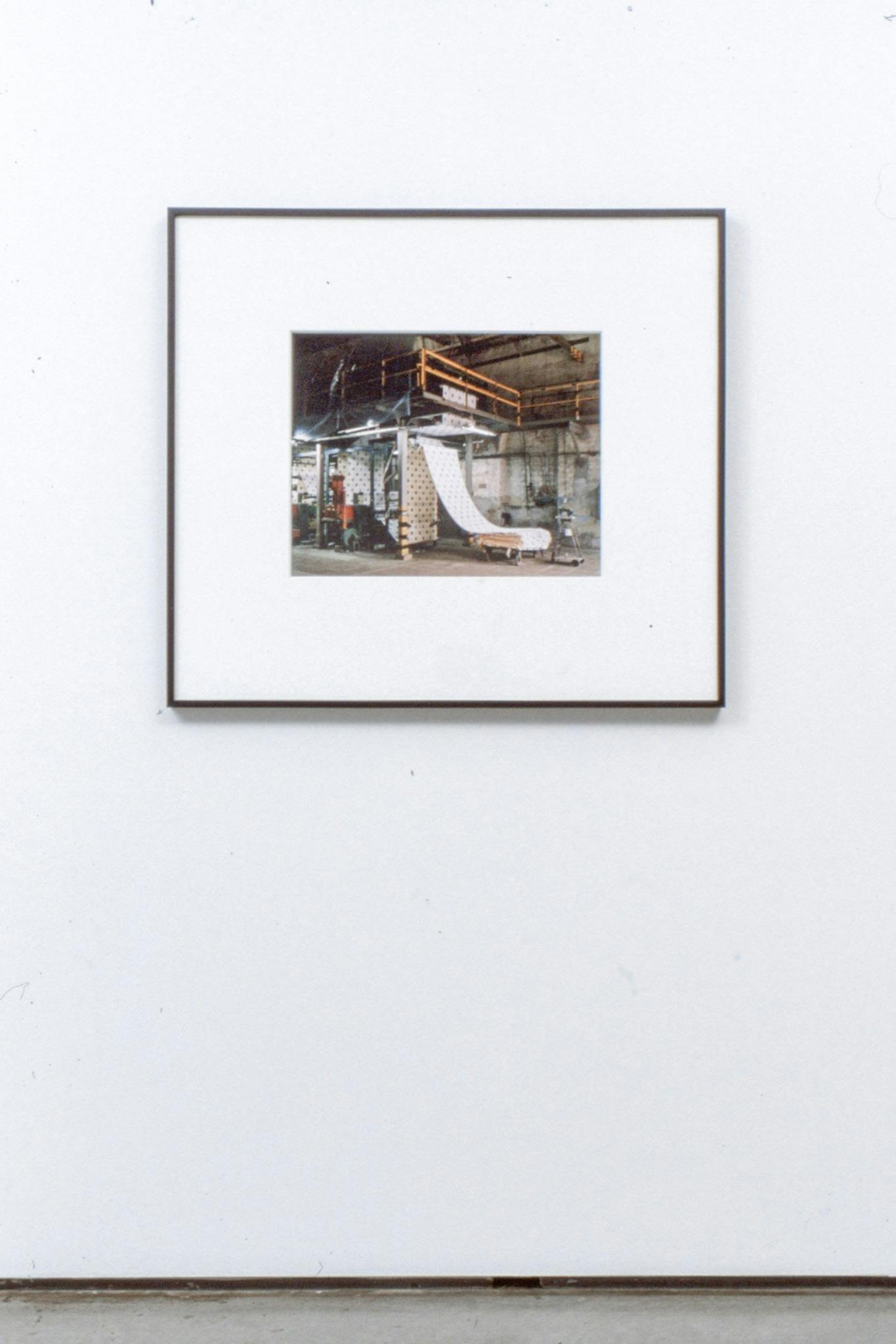 An install image of a photograph on a gallery wall. The photograph captures an industrial scene of machines lined up in a manufacturing firm creating one long sheet of white fabric or paper.