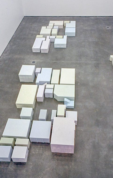 This is an aerial view of the sculptural installation in a gallery space. Pale coloured square sculptures are placed on the floor. They are divided into three groups. 