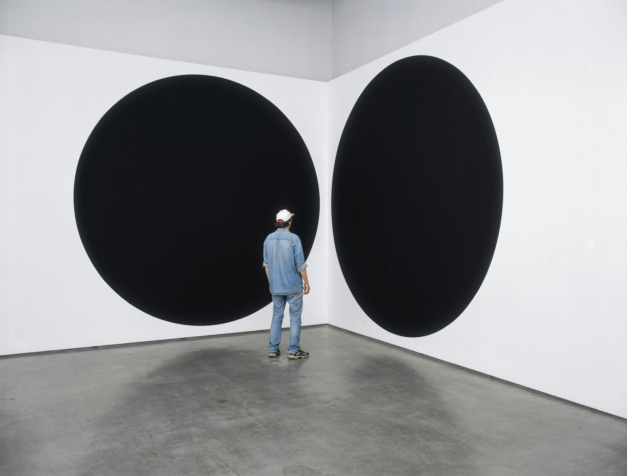 Installation image of paintings by Neil Campbell. The work is a pair of black circles. They are installed on the walls near the corner. The paintings are as tall as the gallery wall, and twice as tall as a person standing in front of them. 