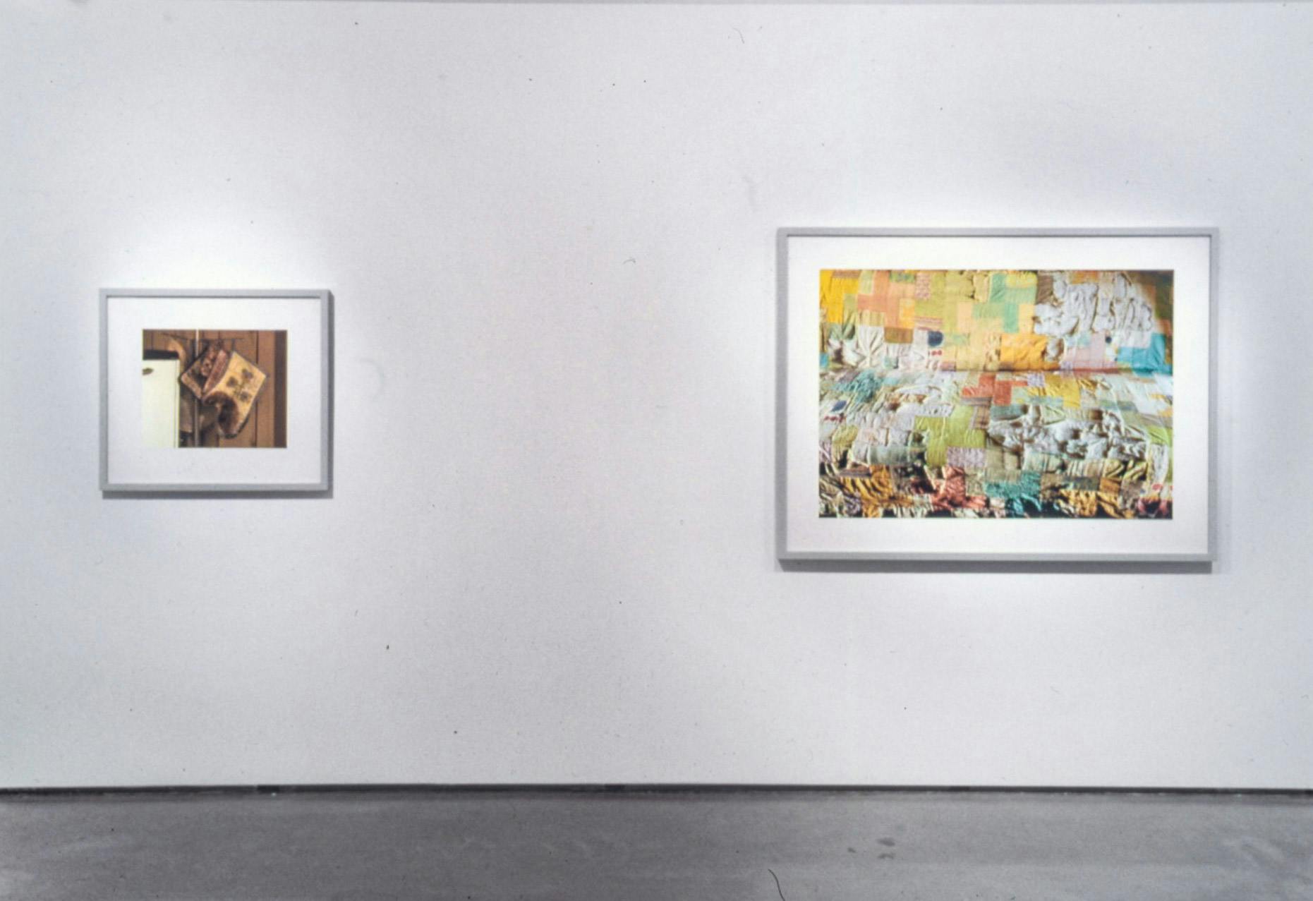 Two photographs are installed on the gallery walls. The photograph on the left depicts cooking mittens. The large photograph on the right depicts a surface of a piece of colourful quilt work. 