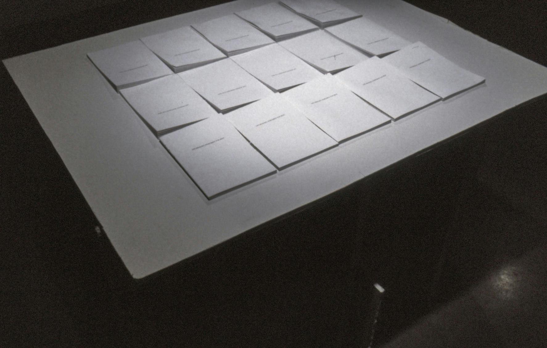 15 sets of paper documents are placed on a white table. The room is dark and the light is casting directly from above the table. 