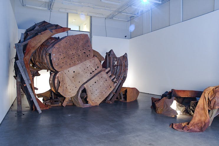 An installation image of large artworks by Donald Lawrence inside a gallery. An assemblage of distorted and decayed metal parts of some large industrial machine is placed on the floor.