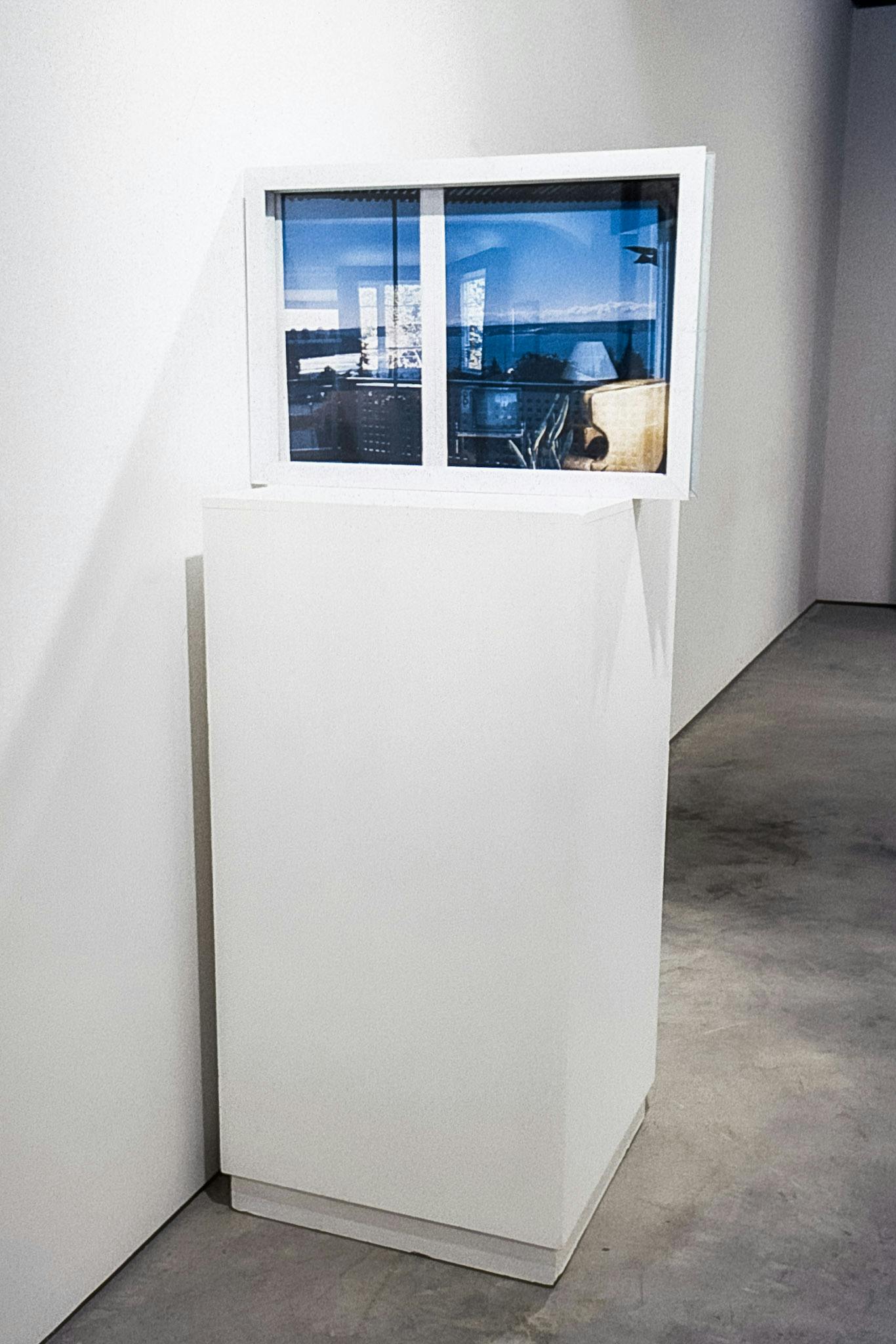 A small glass window with white panes rests on a white plinth against the wall of a gallery. The window appears to reflect a living room with a large window and furniture. 