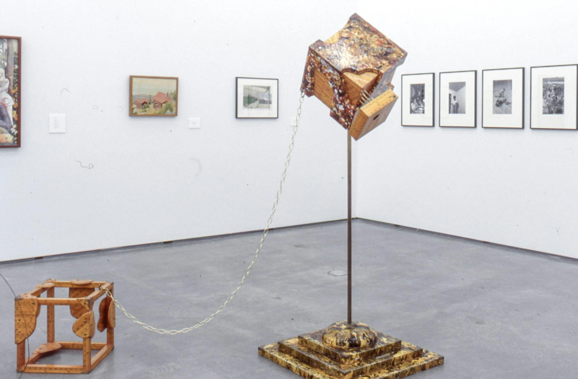 A tall sculpture is placed in the middle of the gallery floor. It has two cube shapes; one is on the floor and another in the air supported by a thin leg. These two cubes are connected with a chain.