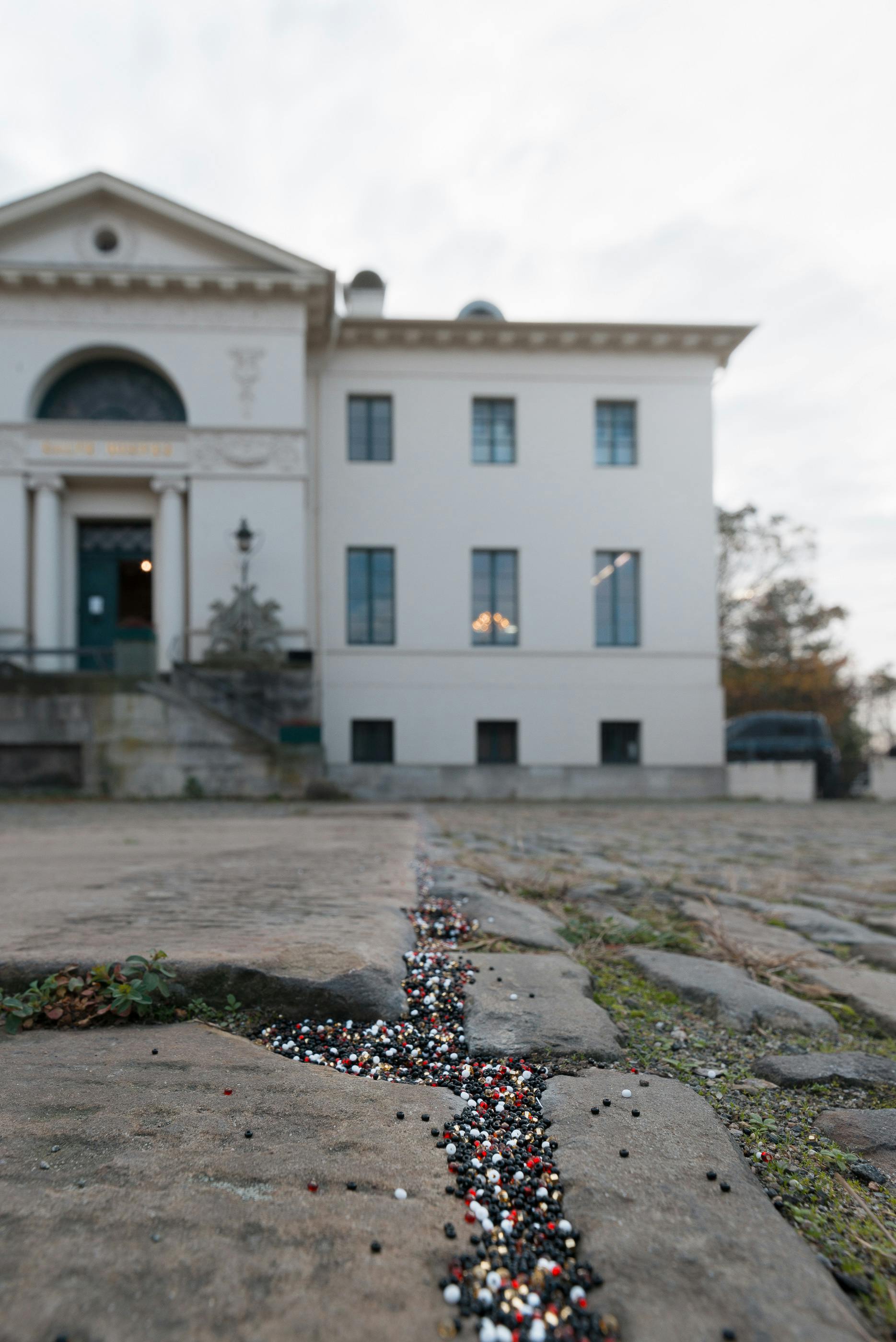 A close view of beads black, red, white, and gold beads filling the cracks of cobblestone pavement. Part of a white building is blurred in the background.