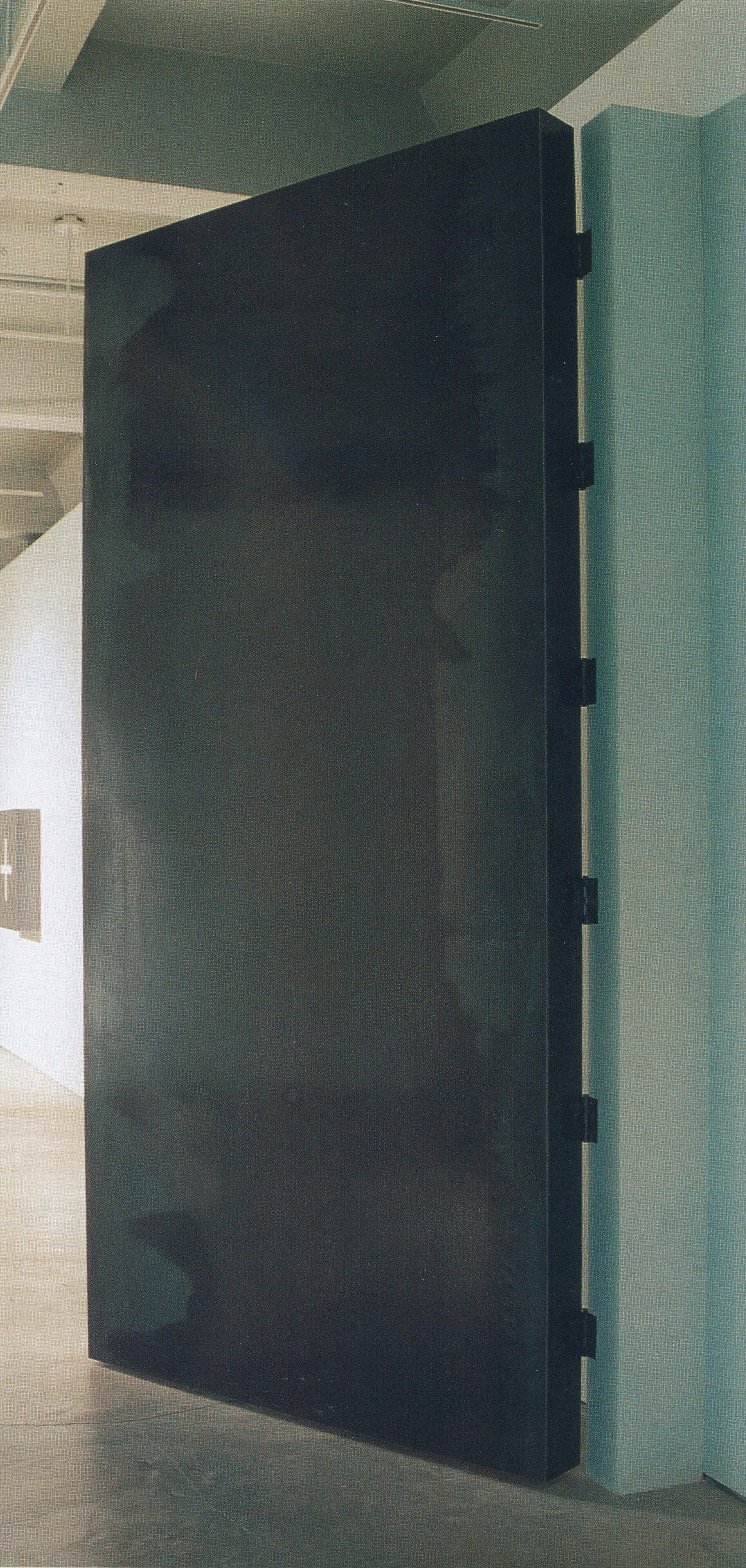 A large steel-made artwork is installed in a gallery. It’s overall colour is black, and some oil-stain patterns appear on the surface. It is door-like shaped and attached to a gallery wall with hinges. 