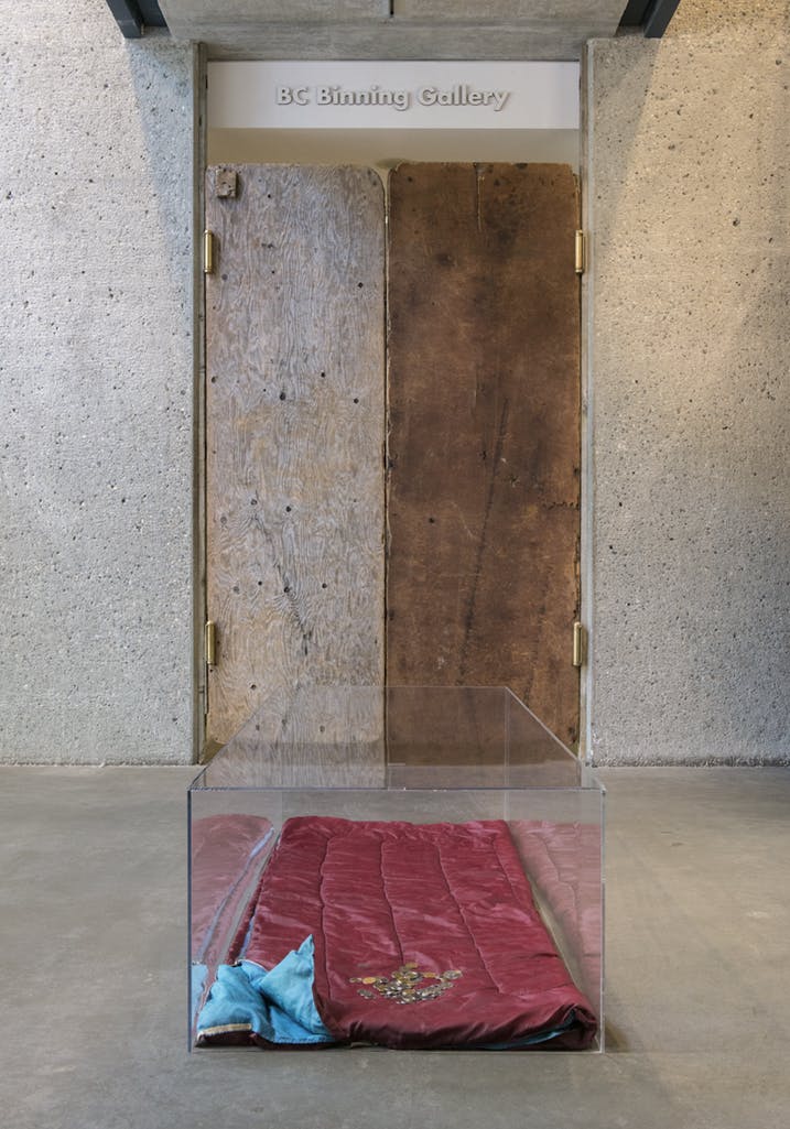 A red sleeping bag lies on the floor of a lobby under a display case cover. Coins are placed on one edge of the sleeping bag. Behind, wooden double doors are installed at the entrance to a gallery.