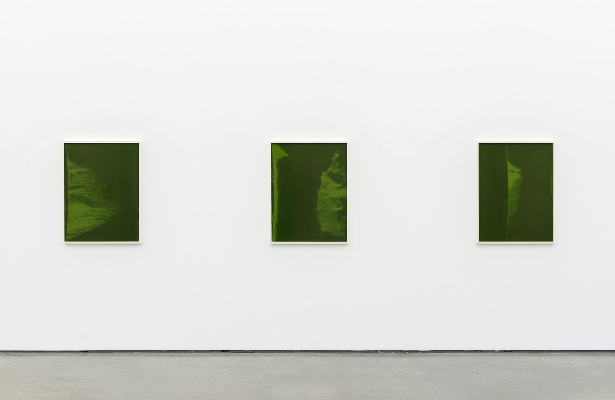 Three framed photographic prints hang on the wall of a gallery space. The images are of the surface of the sun overlaid with the frame’s deep, green glass. 